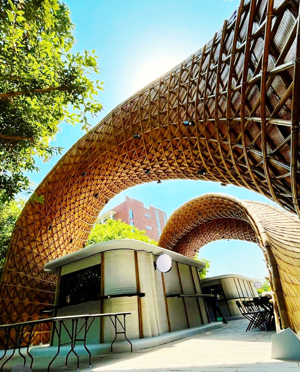 Curved, patterned wooden arches over modern outdoor kiosks with tables under a bright blue sky.