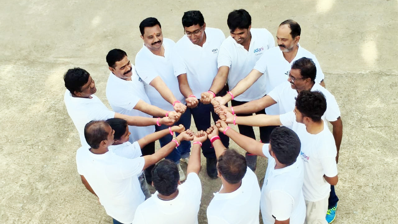 A group of men in white Adani shirts and pink wristbands stand in a circle, touching fists in unity.