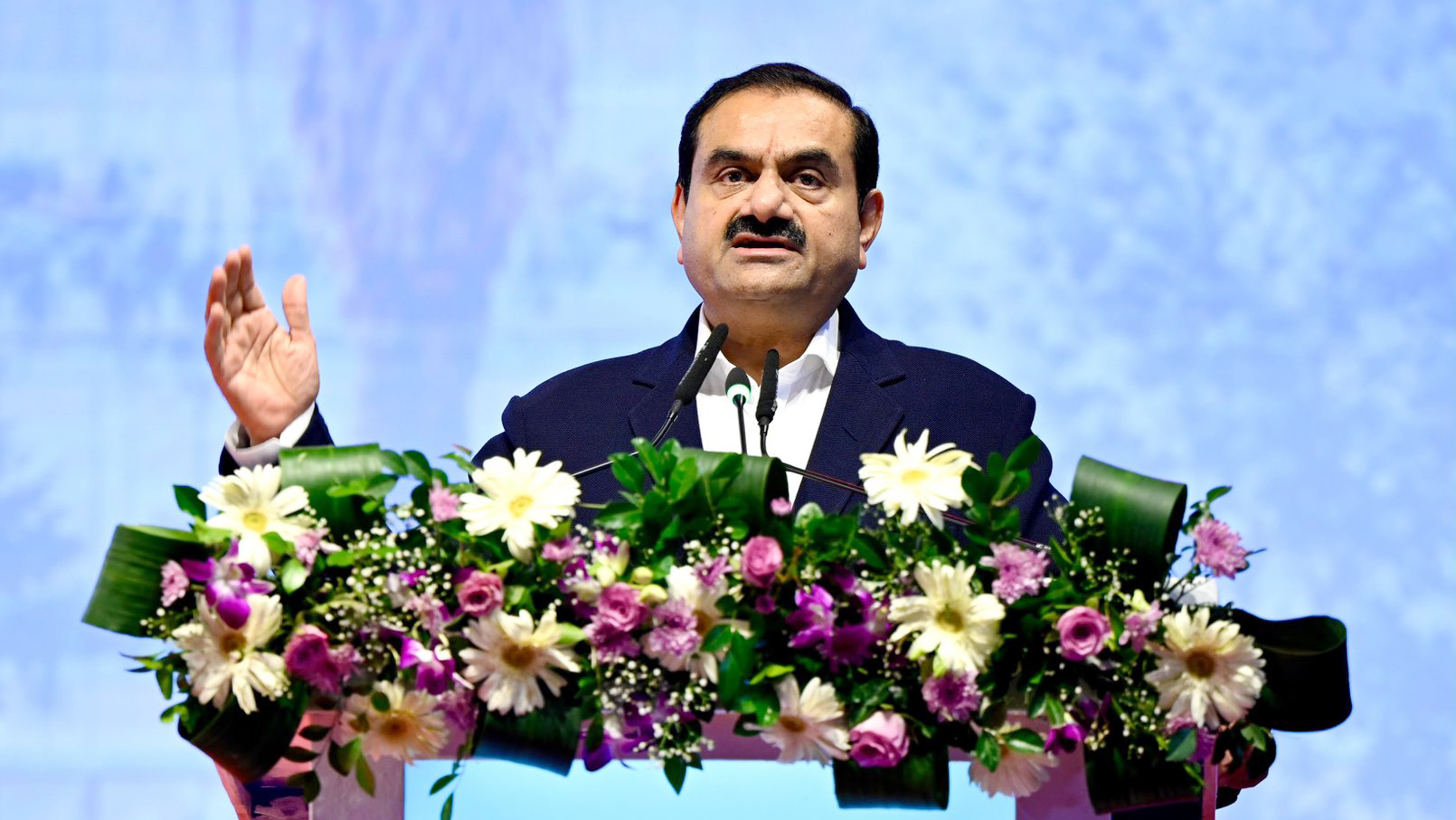 Gautam Adani speaks at a podium adorned with flowers, raising his left hand.