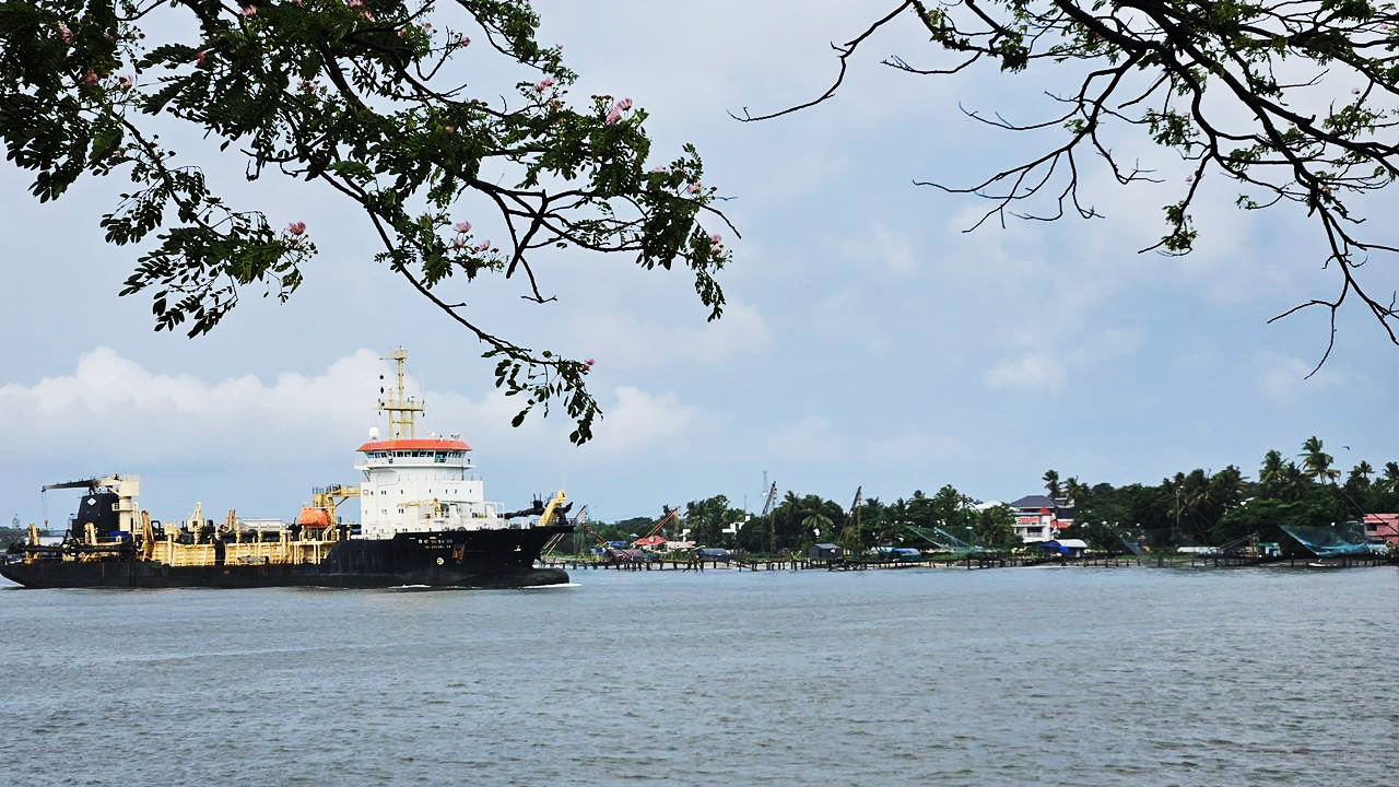 A large dredger ship moves on a wide river, framed by flowering branches, with a village in the background.
