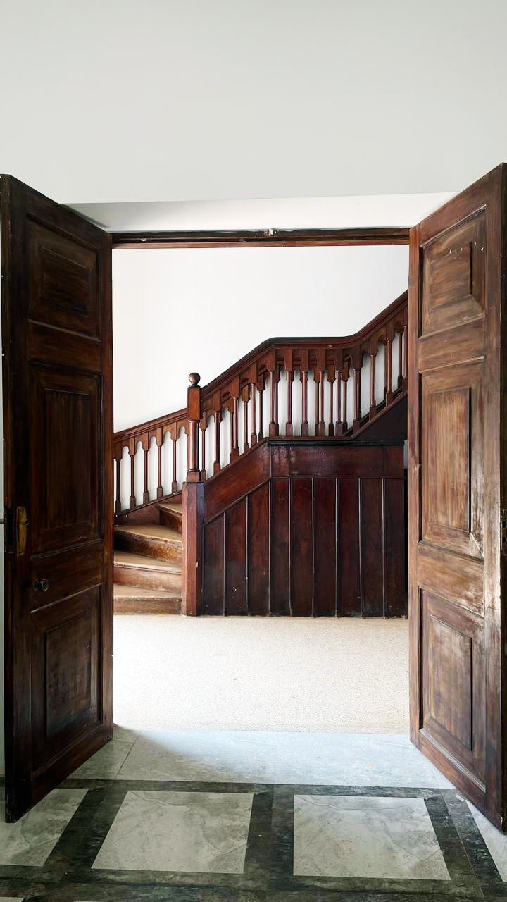 A view through open ornate dark wooden doors revealing an elegant curved wooden staircase with white walls.