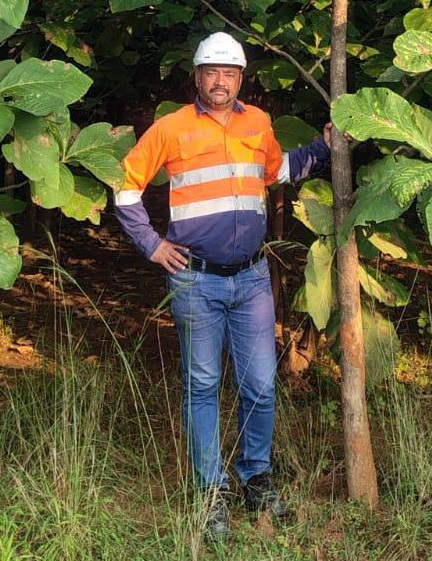 A man in a white hard hat and high-vis shirt stands by a tree in a lush green outdoor setting.