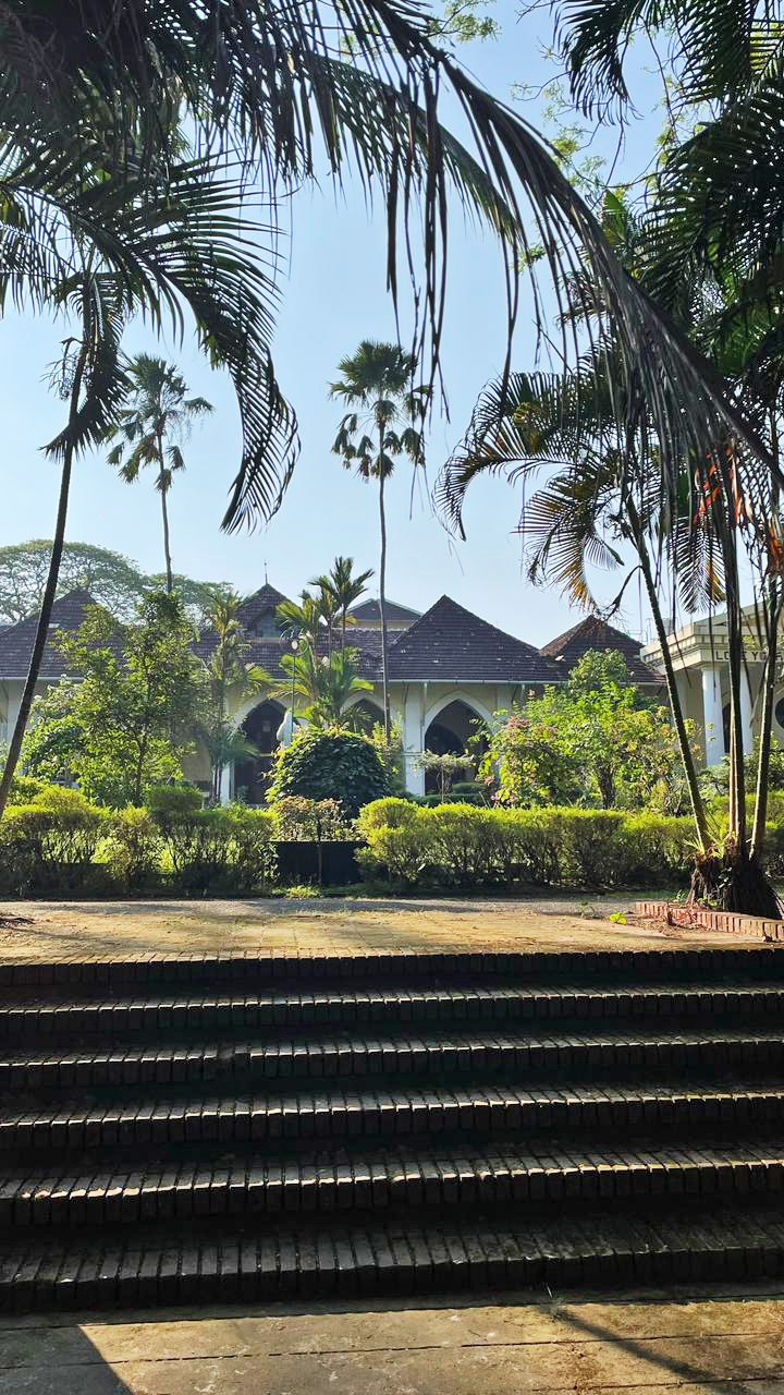Ornate tropical building with tiled roof, lush gardens, palm trees, and foreground brick steps under a clear sky.