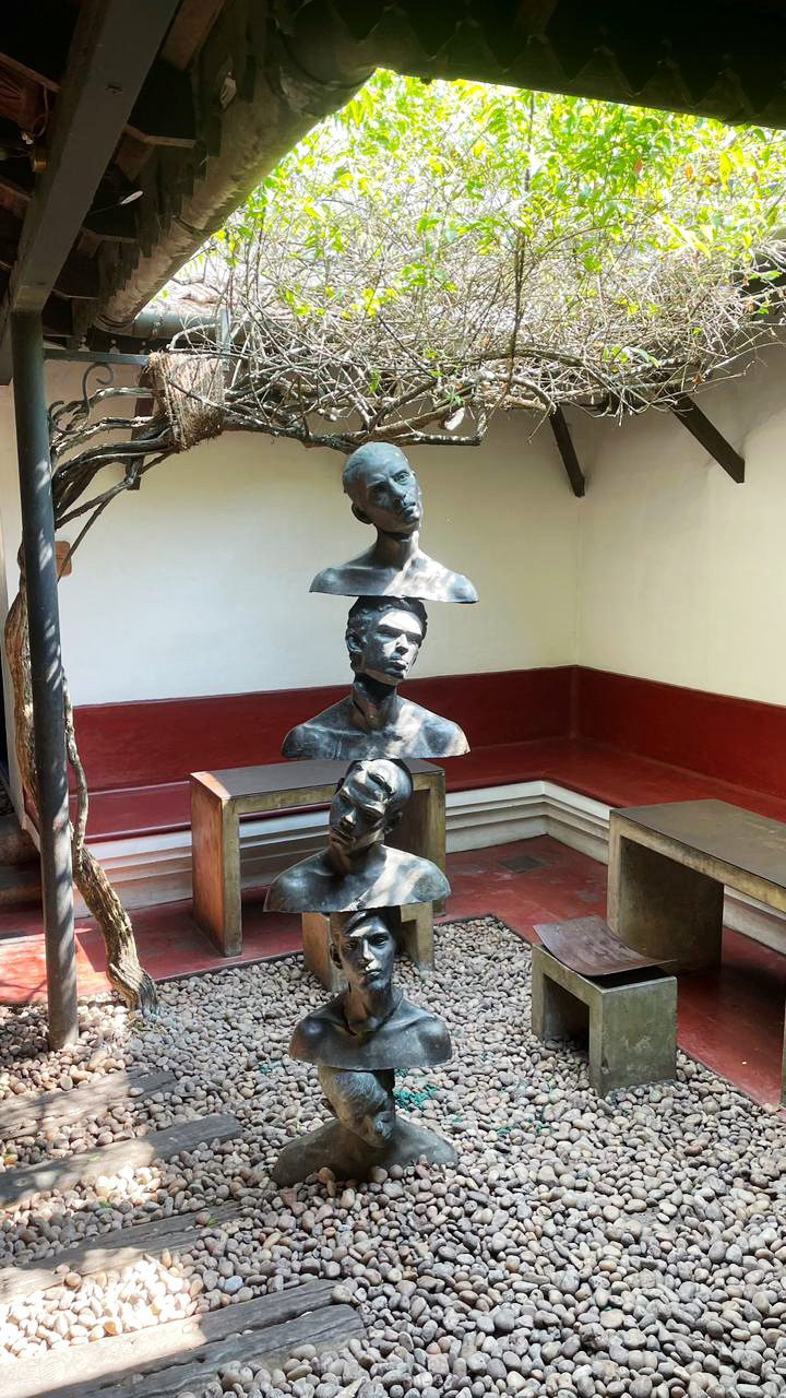 Five bronze busts stacked vertically in a pebbled outdoor courtyard with lush greenery overhead.
