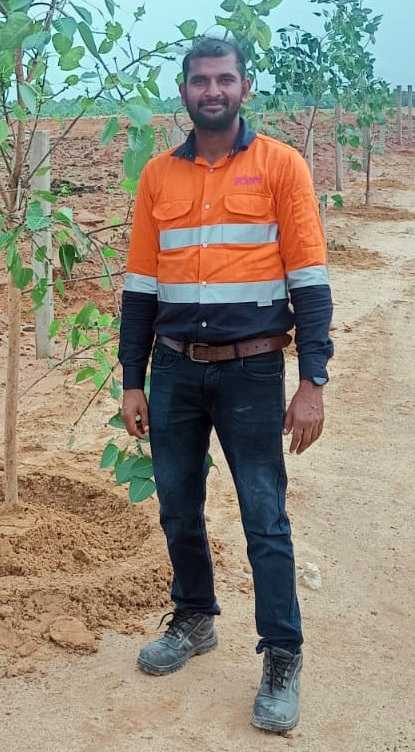 Man in orange and blue work shirt and jeans standing in a field with saplings.