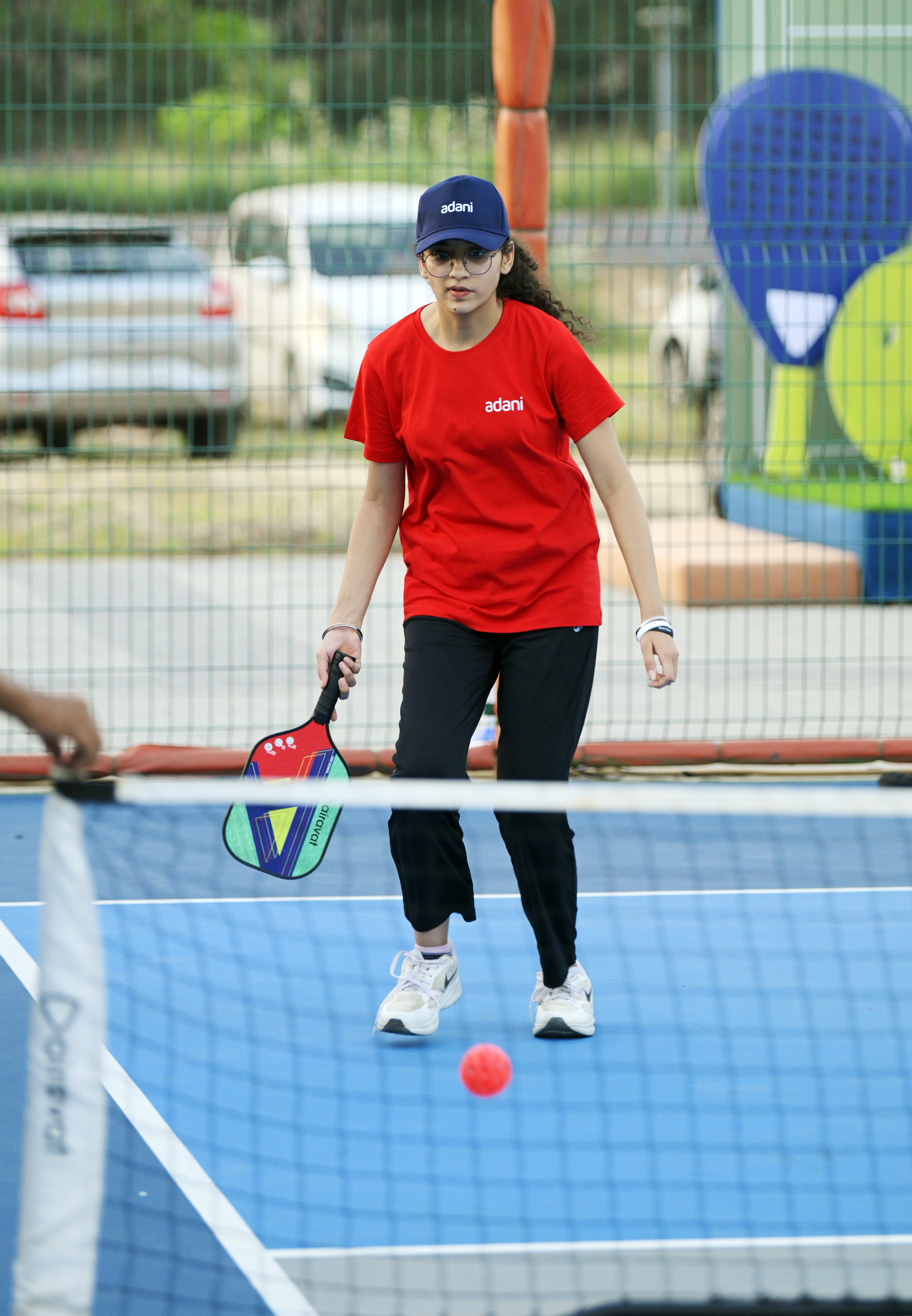 Young girl in red Adani shirt and blue cap playing pickleball on a blue court with a red ball.