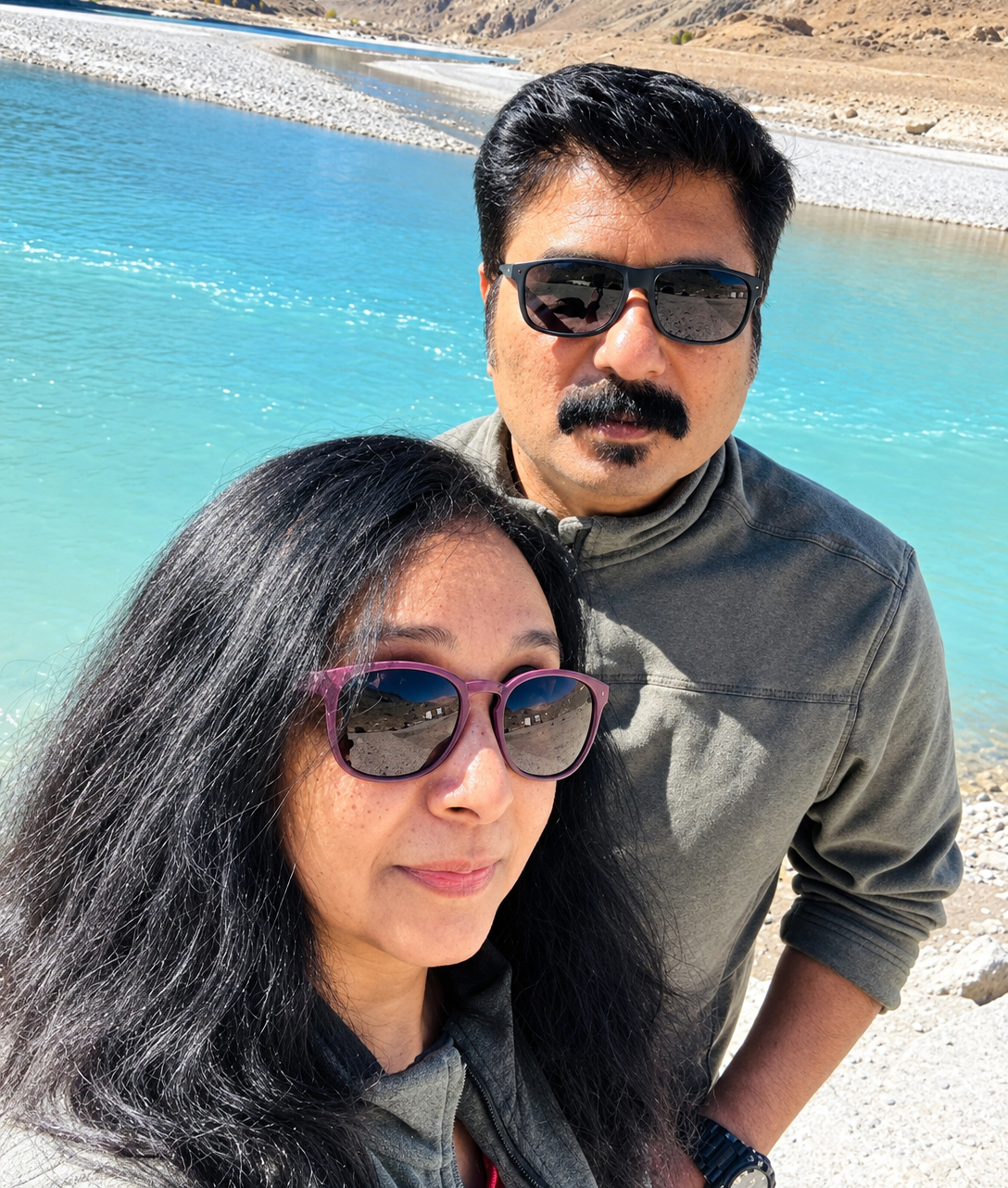 A couple poses for a selfie by a bright turquoise river with mountains in the background under a sunny sky.