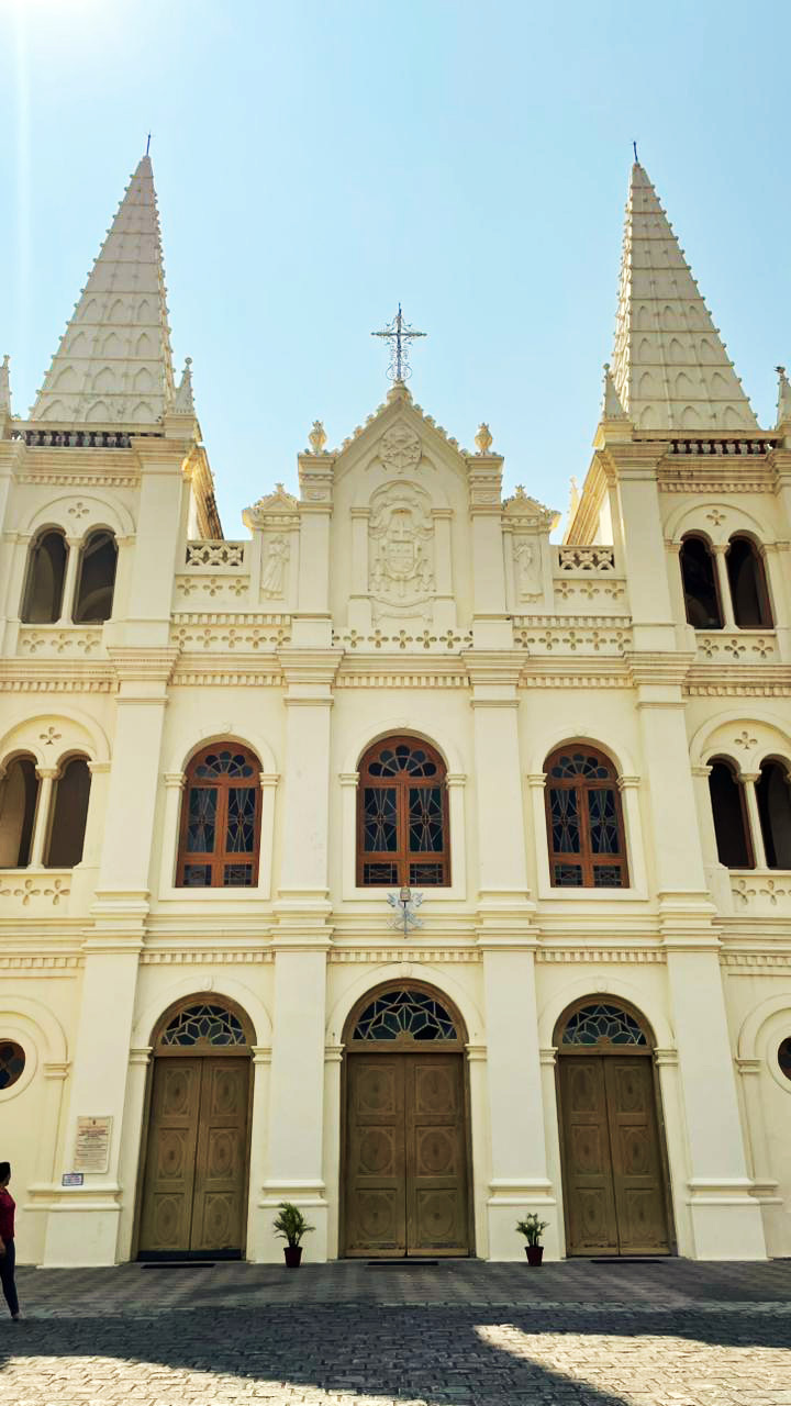 A grand cream-colored church facade with twin spires, arched windows, and three doors under a clear blue sky.