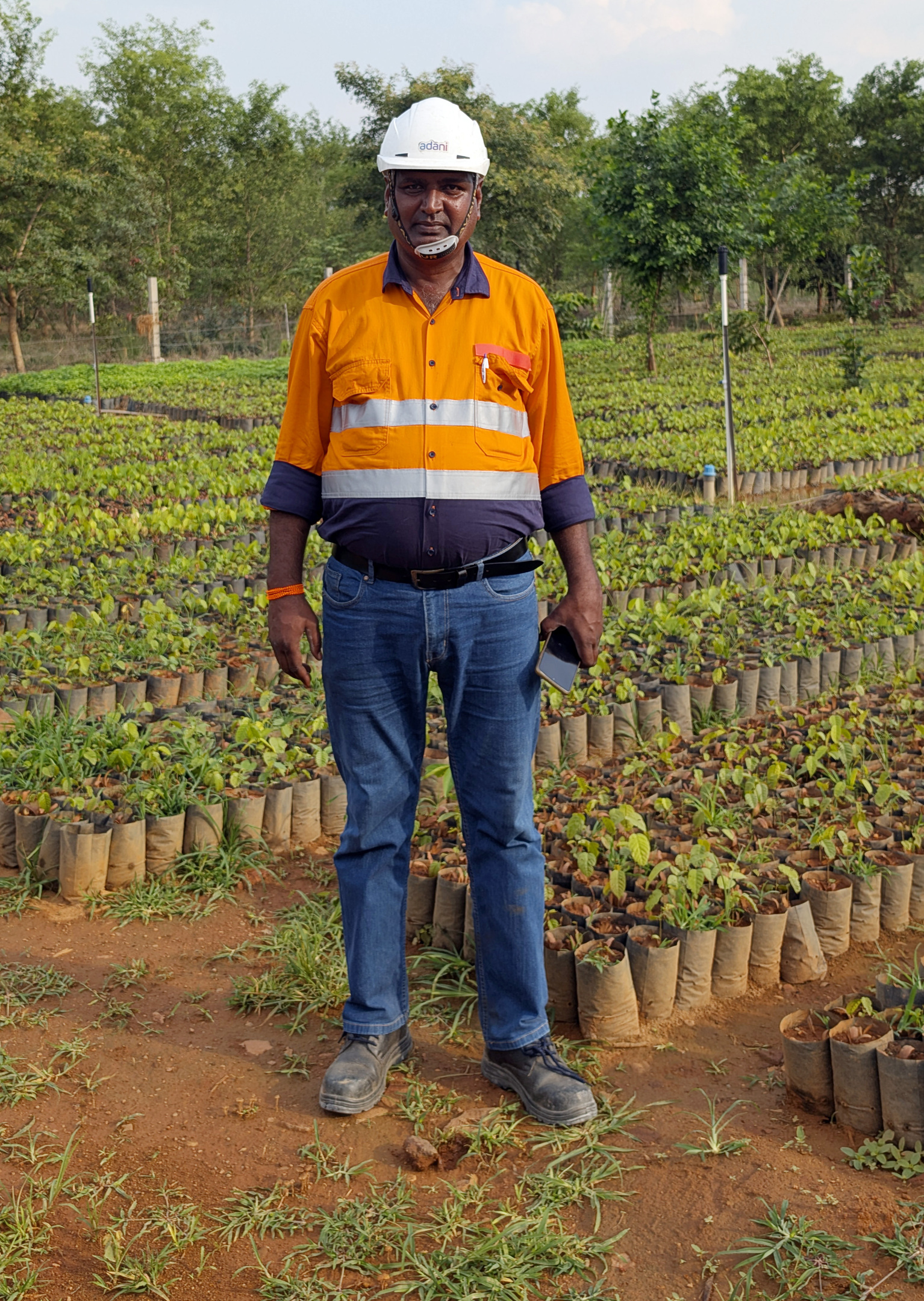 A man in an orange and blue work shirt, jeans, and a white hard hat stands in a nursery full of small plants.