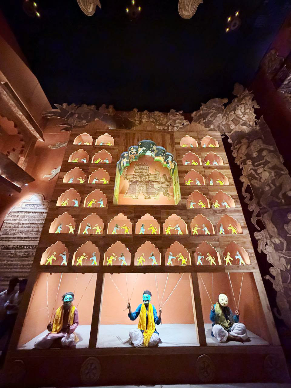 An elaborate Ramayana-themed puppet display with three masked puppeteers and tiered niches showing scenes.