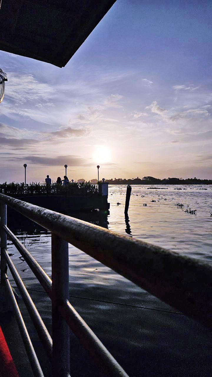 A boat on a serene lake during sunset, with a metal railing in the foreground and people on a distant dock.