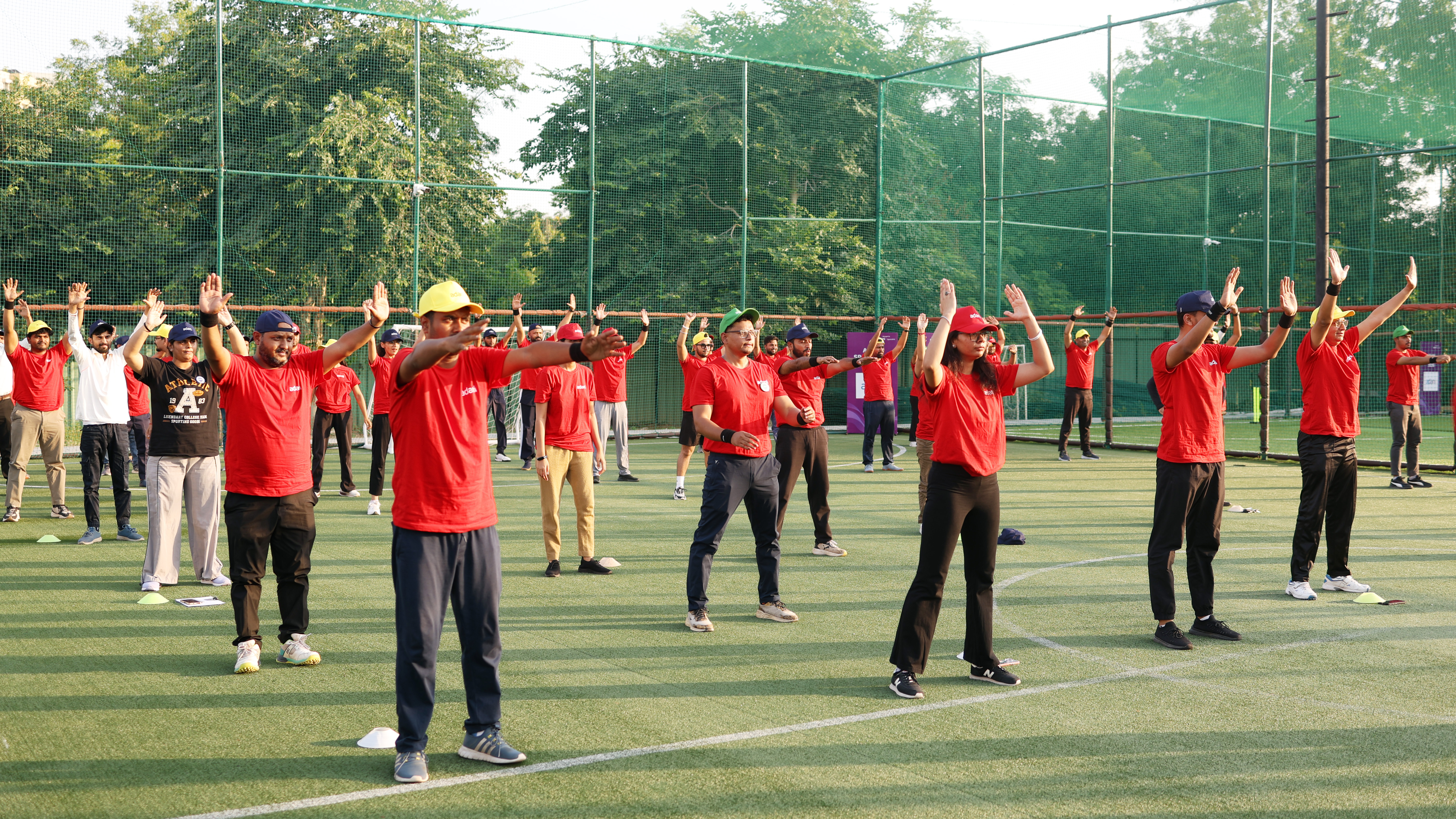 A group of people in red shirts and hats perform outdoor arm stretches on a green artificial turf field.