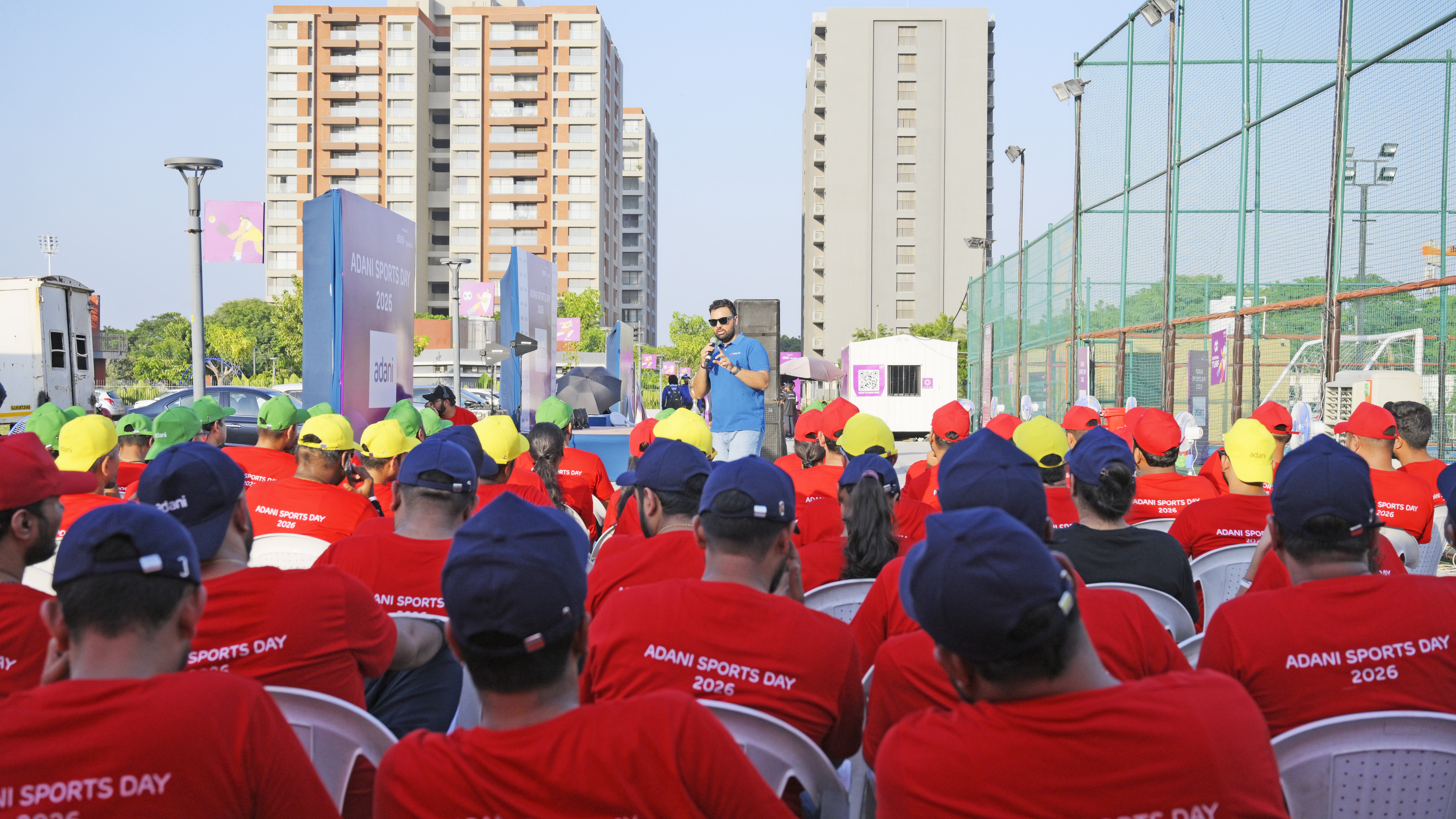 Man speaks to audience wearing 'Adani Sports Day 2026' shirts at an outdoor event.