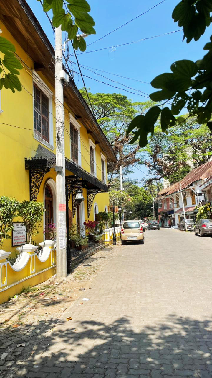 A vibrant yellow building lines a sunlit street in Goa, with cars and lush trees.