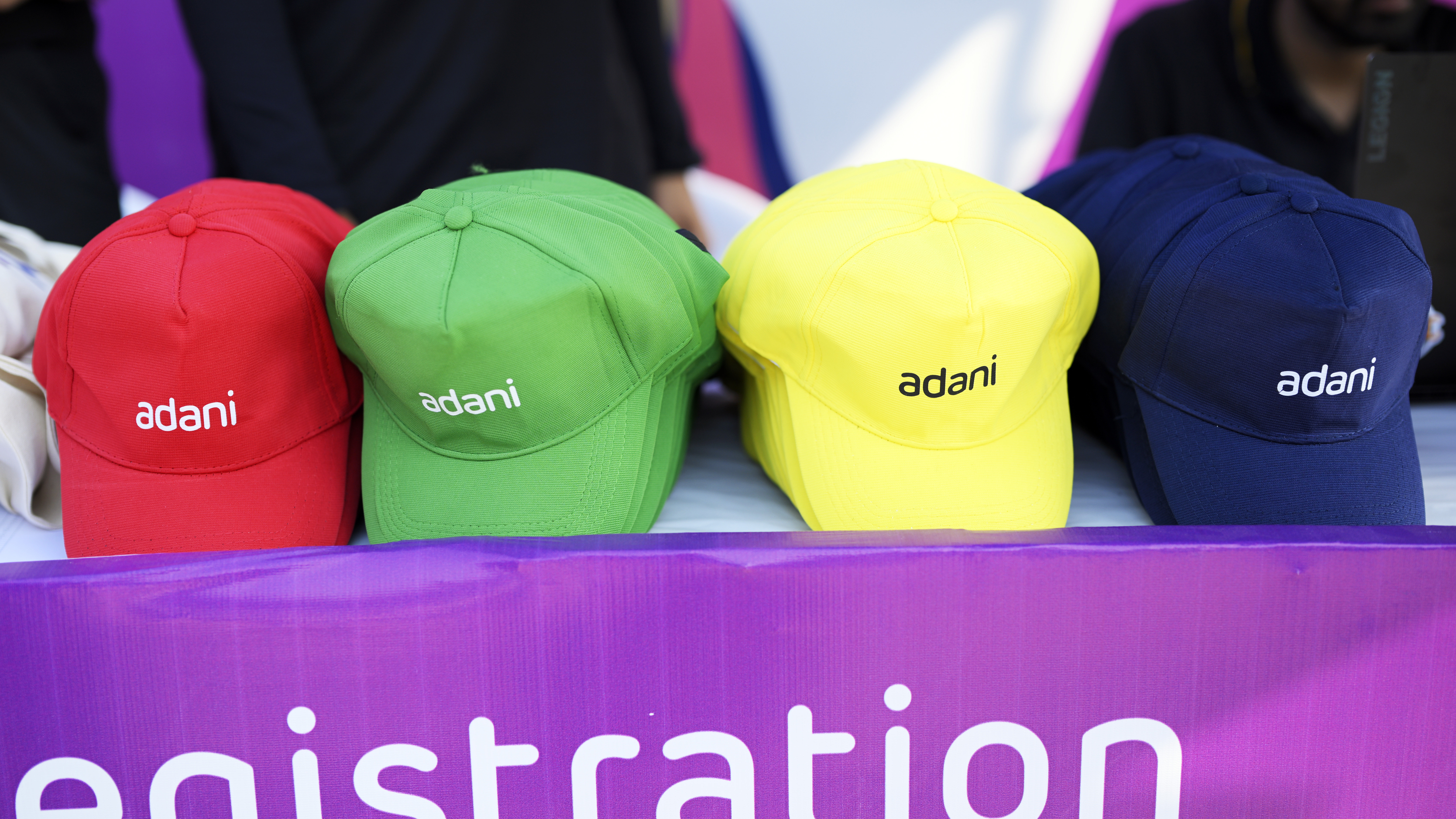 Four colorful "adani" baseball caps (red, green, yellow, blue) on a table with a partially visible "registration" banner.