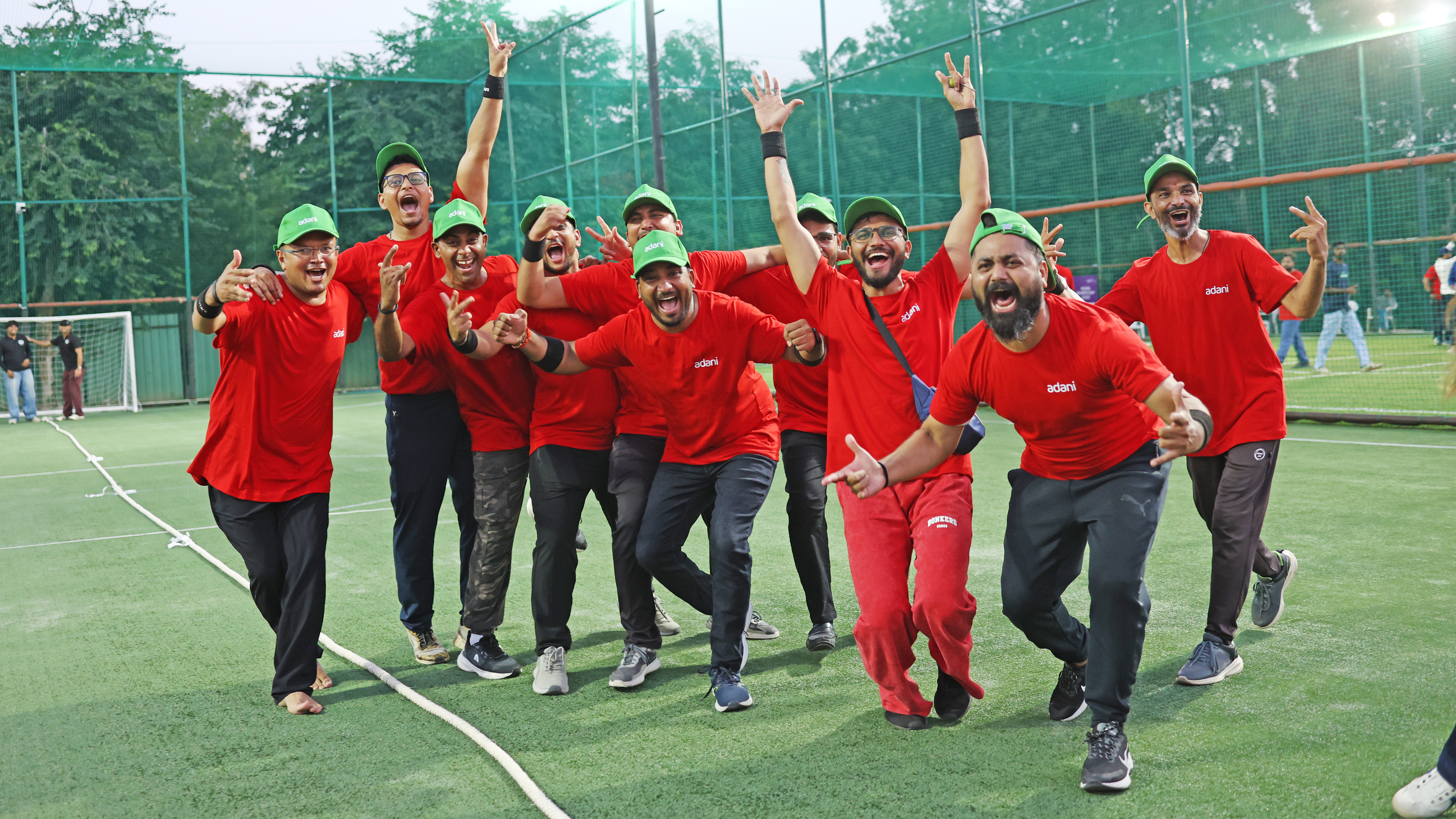 A group of men in red 'adani' t-shirts and green caps celebrating vigorously on a green sports field.