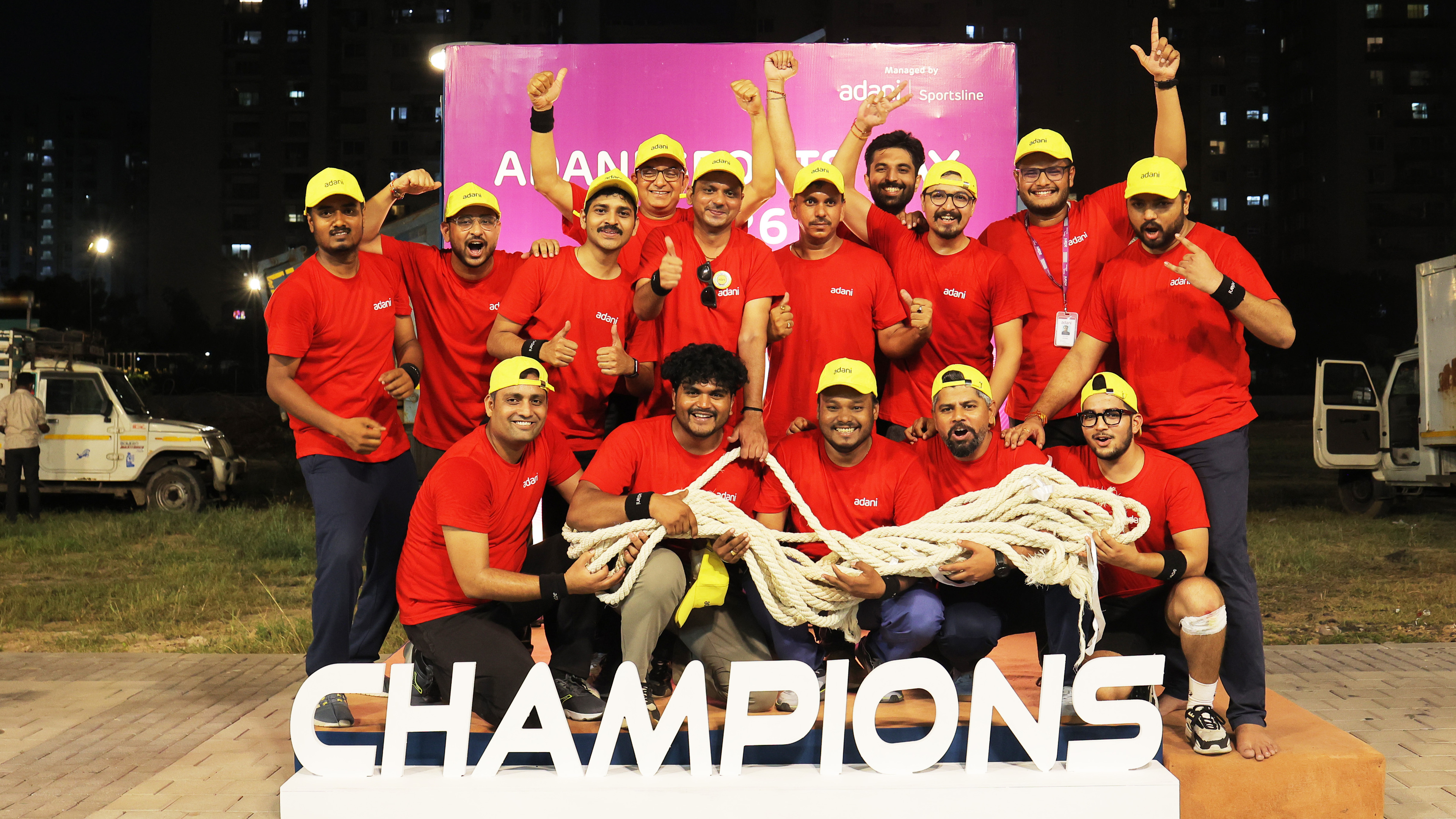 A large group of men in red shirts and yellow caps celebrate with a rope and a "CHAMPIONS" sign.