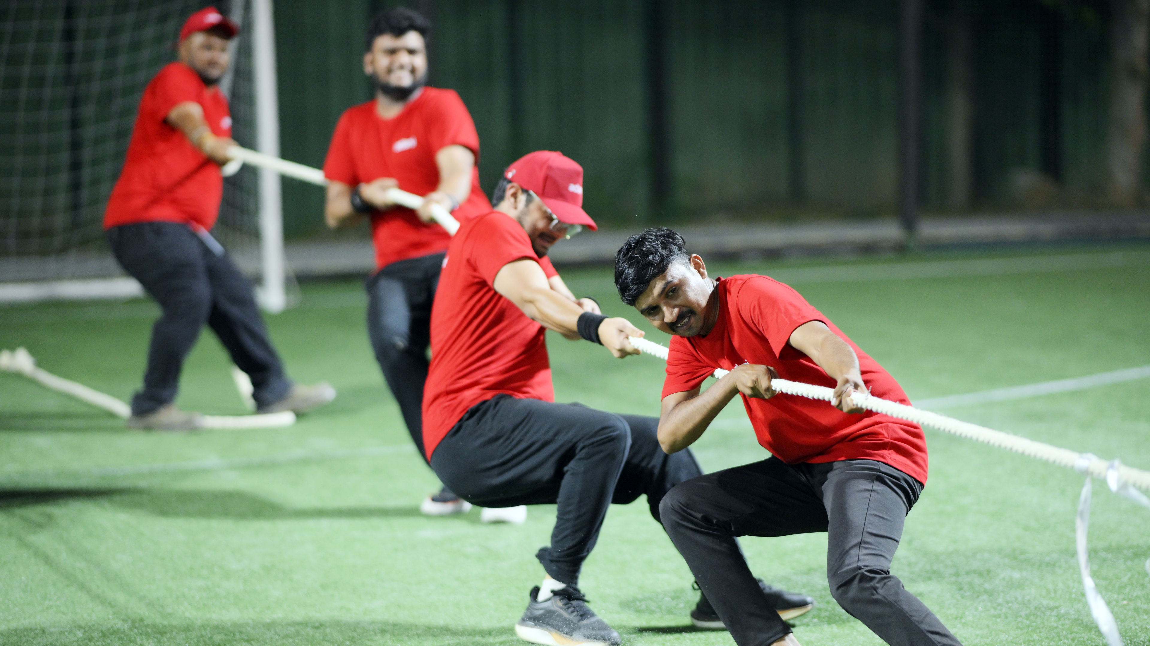 Four men in red shirts playing a spirited game of tug of war on a green artificial turf.