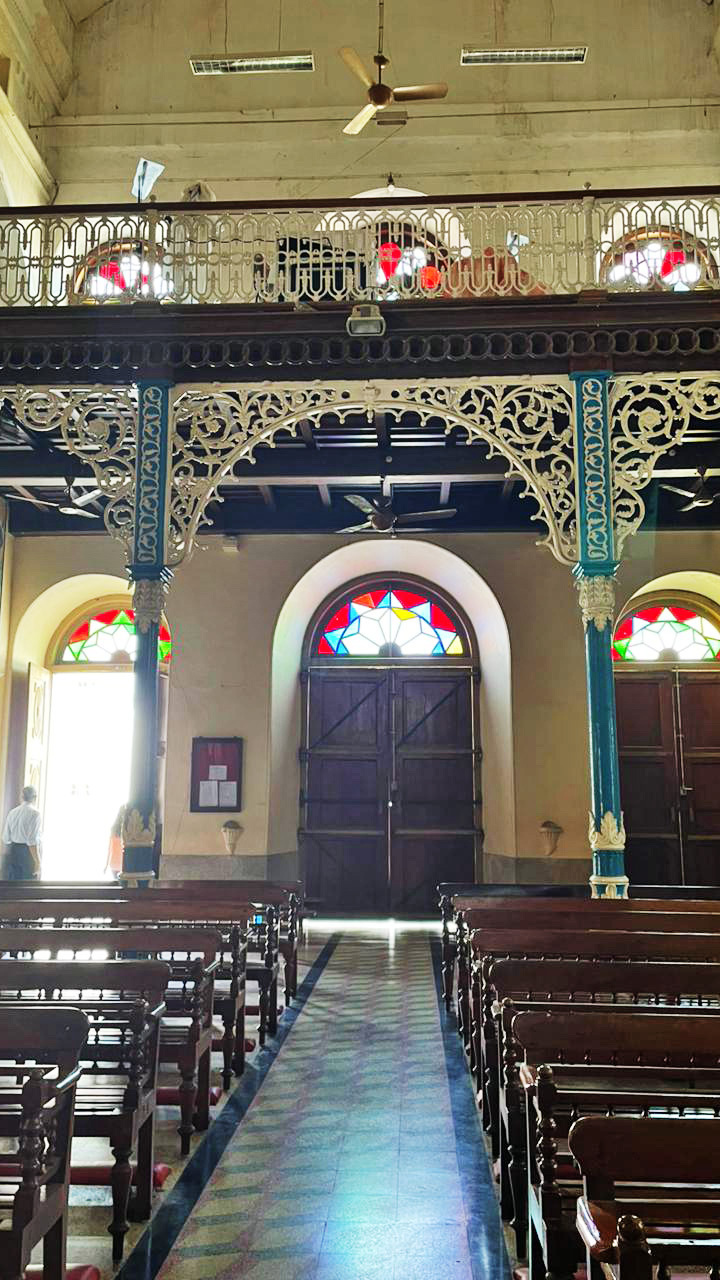 View of a church or old building interior with a central aisle, wooden pews, stained glass, and an ornate white and blue balcony.