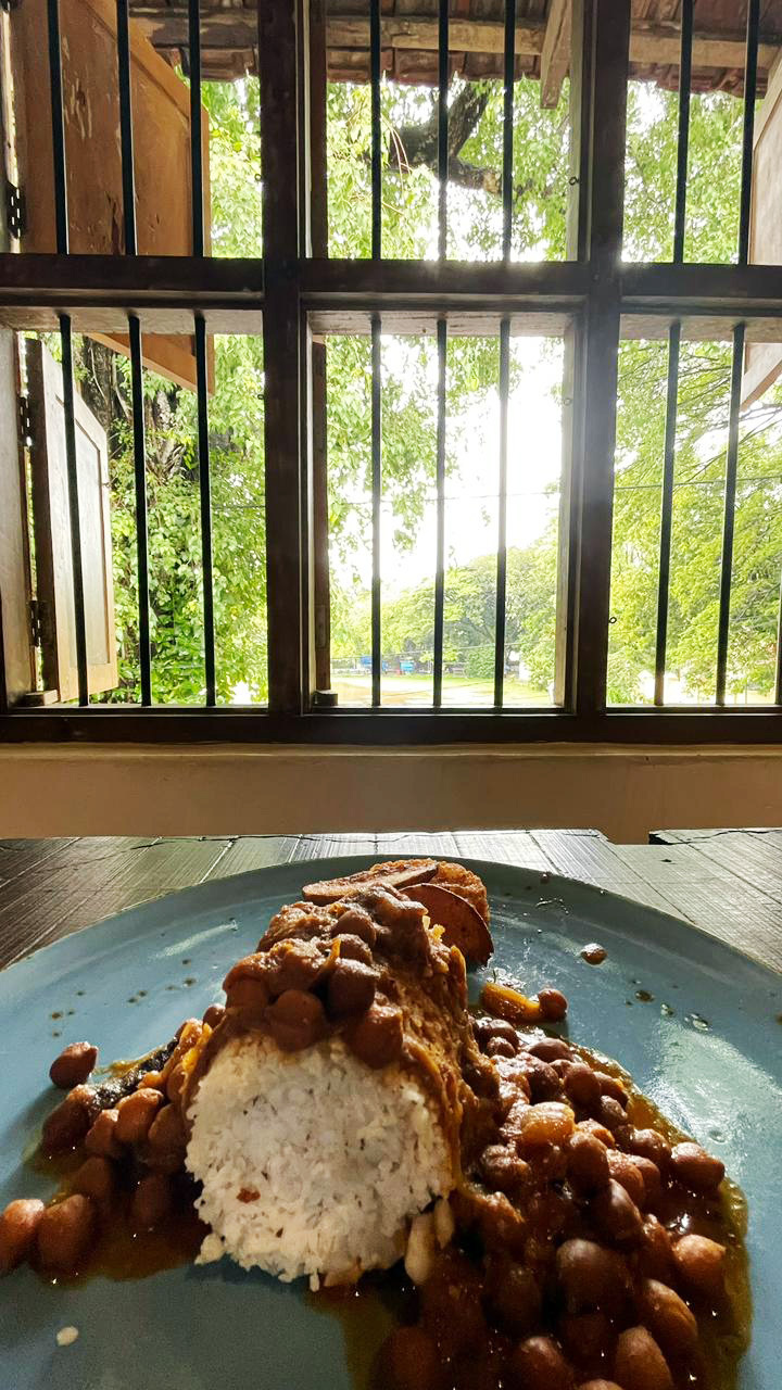 A dish of rice and bean stew on a table, with a window featuring bars and a view of green trees outside.