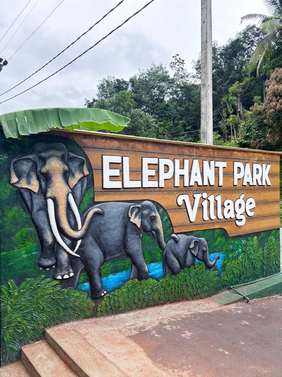 A sign for "ELEPHANT PARK Village" featuring sculpted elephants and lush greenery.