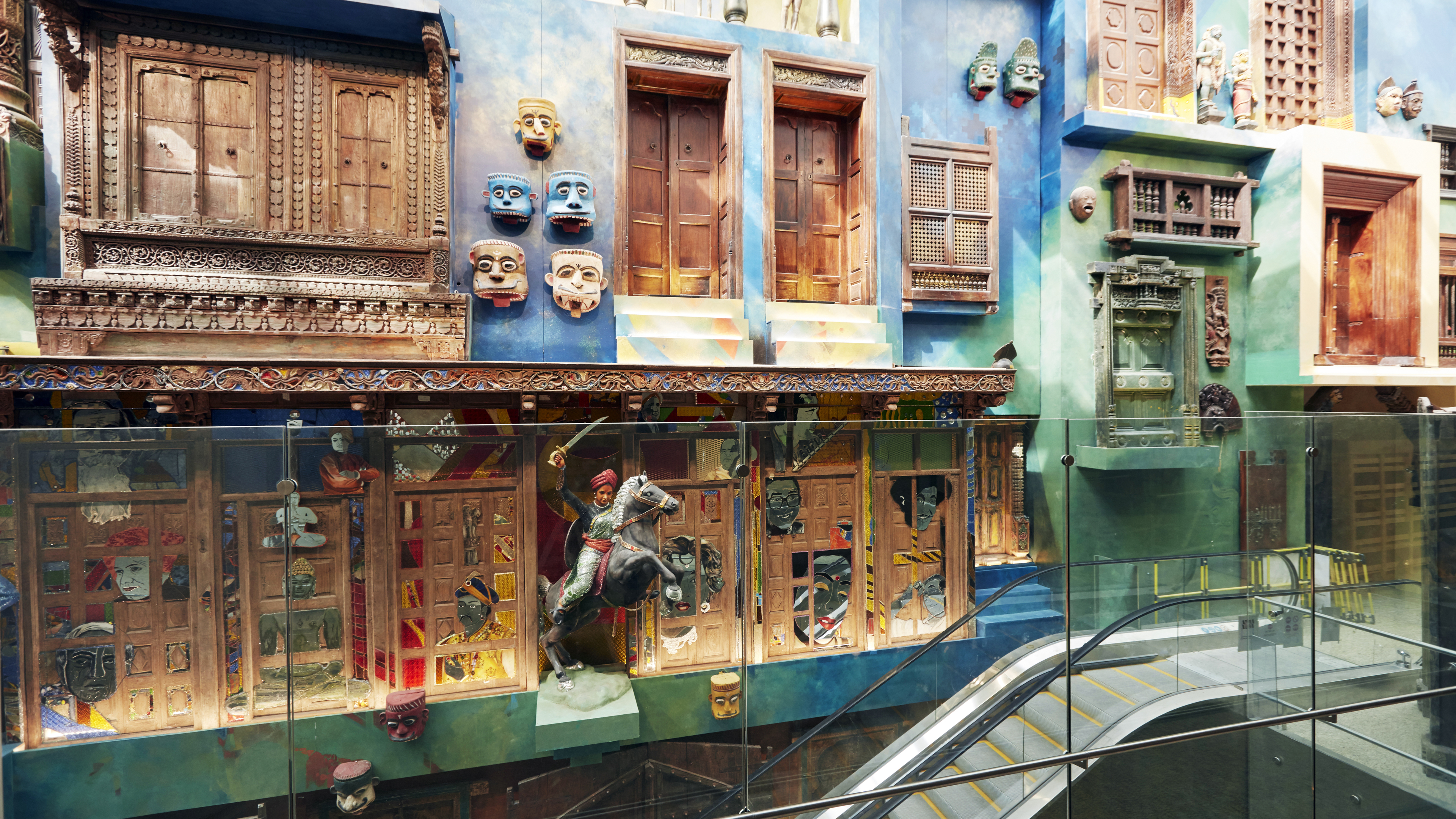 Colorful architectural display featuring carved wooden doors, windows, masks, and a statue, with an escalator.