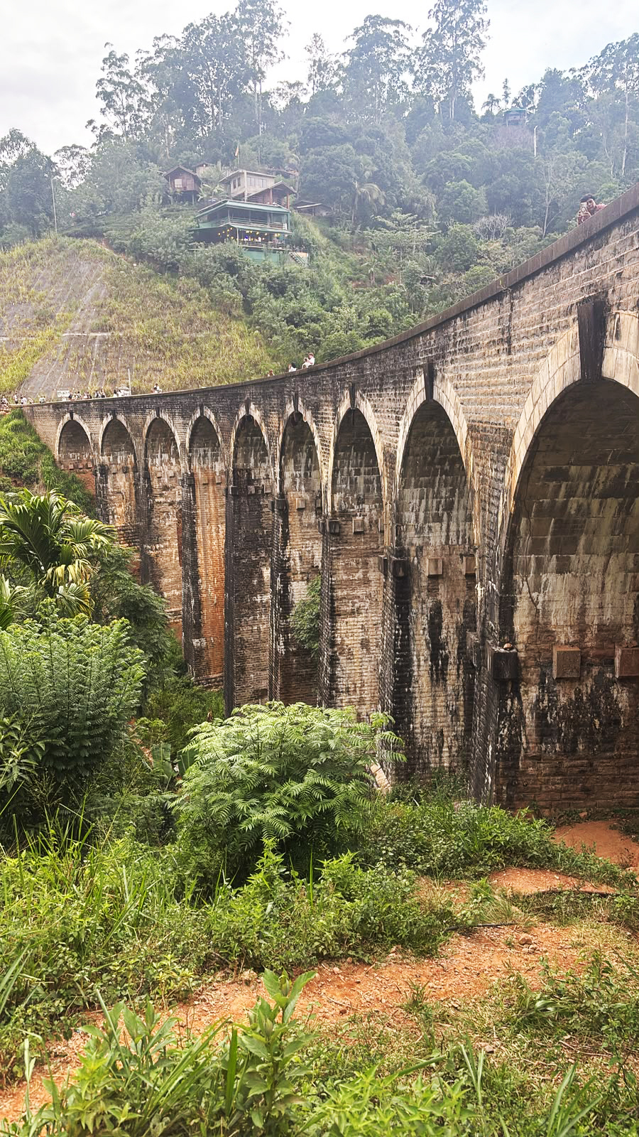 A historic stone viaduct with nine arches, surrounded by lush green foliage and hills with small houses.