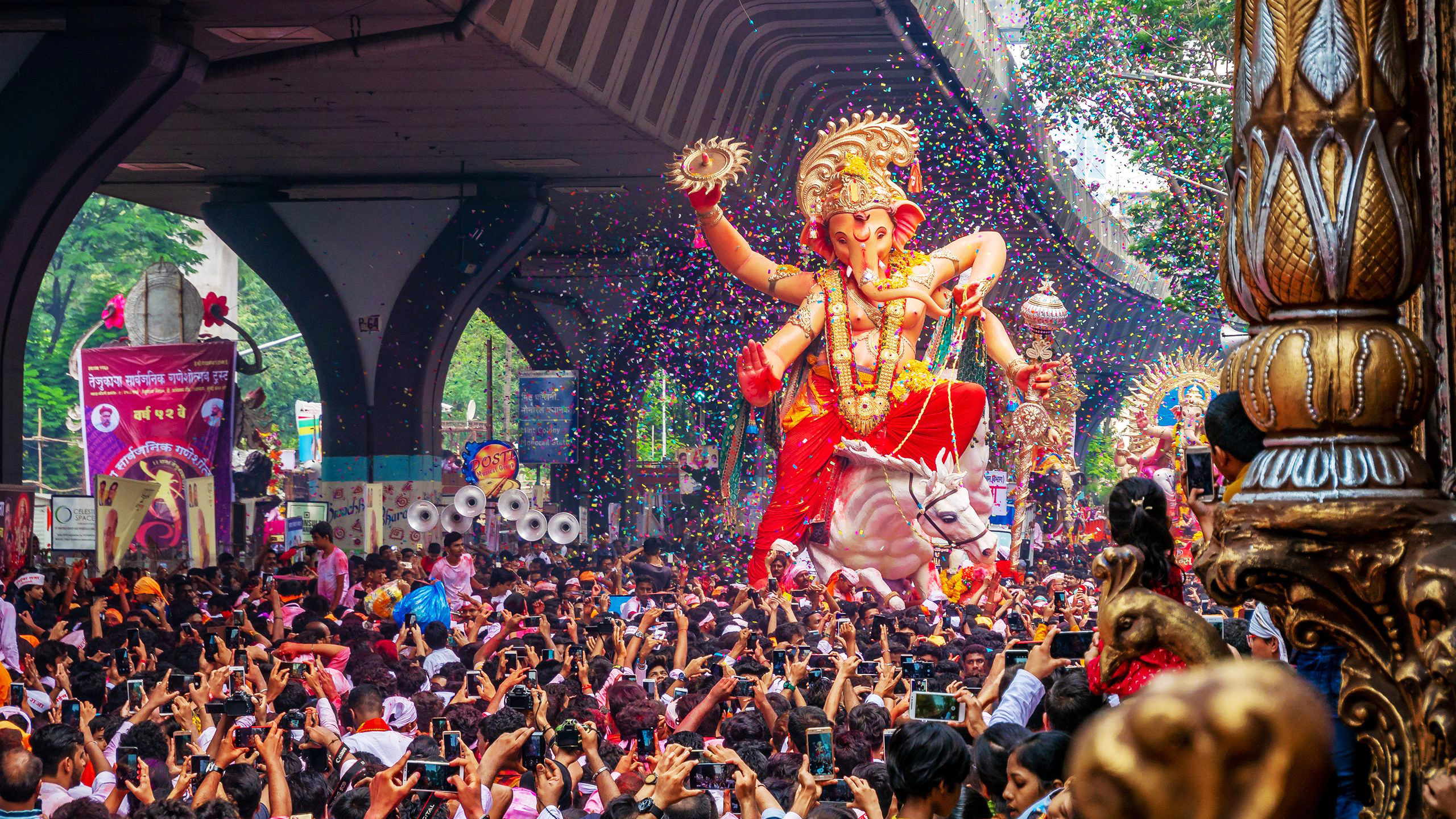 A large Ganesha idol on a float in a vibrant festival procession with confetti and a huge crowd.