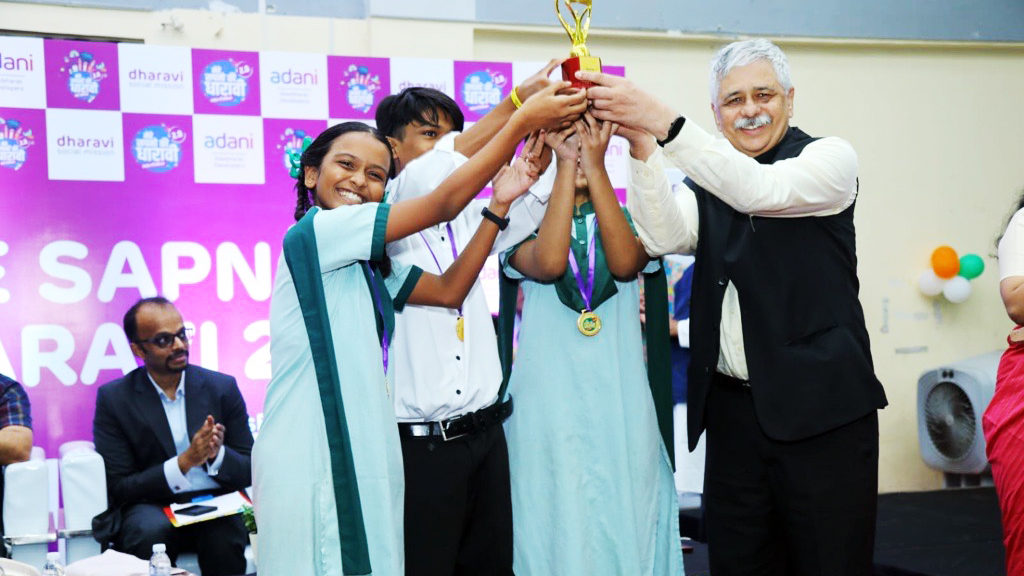 A man and three students smile while holding a golden trophy together on stage at an awards ceremony.