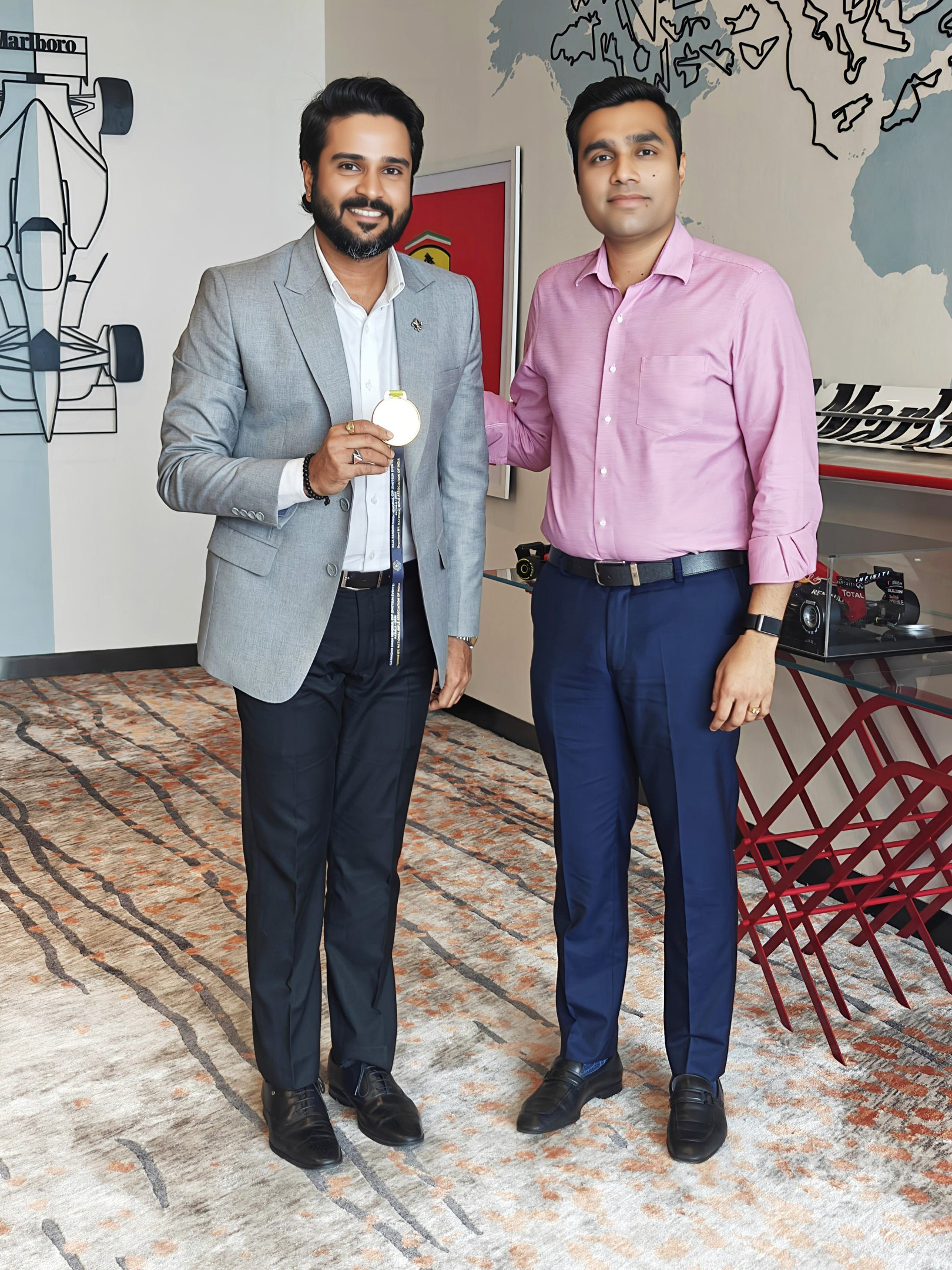 Two men, one holding a gold medal, stand smiling in a room with car-themed decor on the walls.