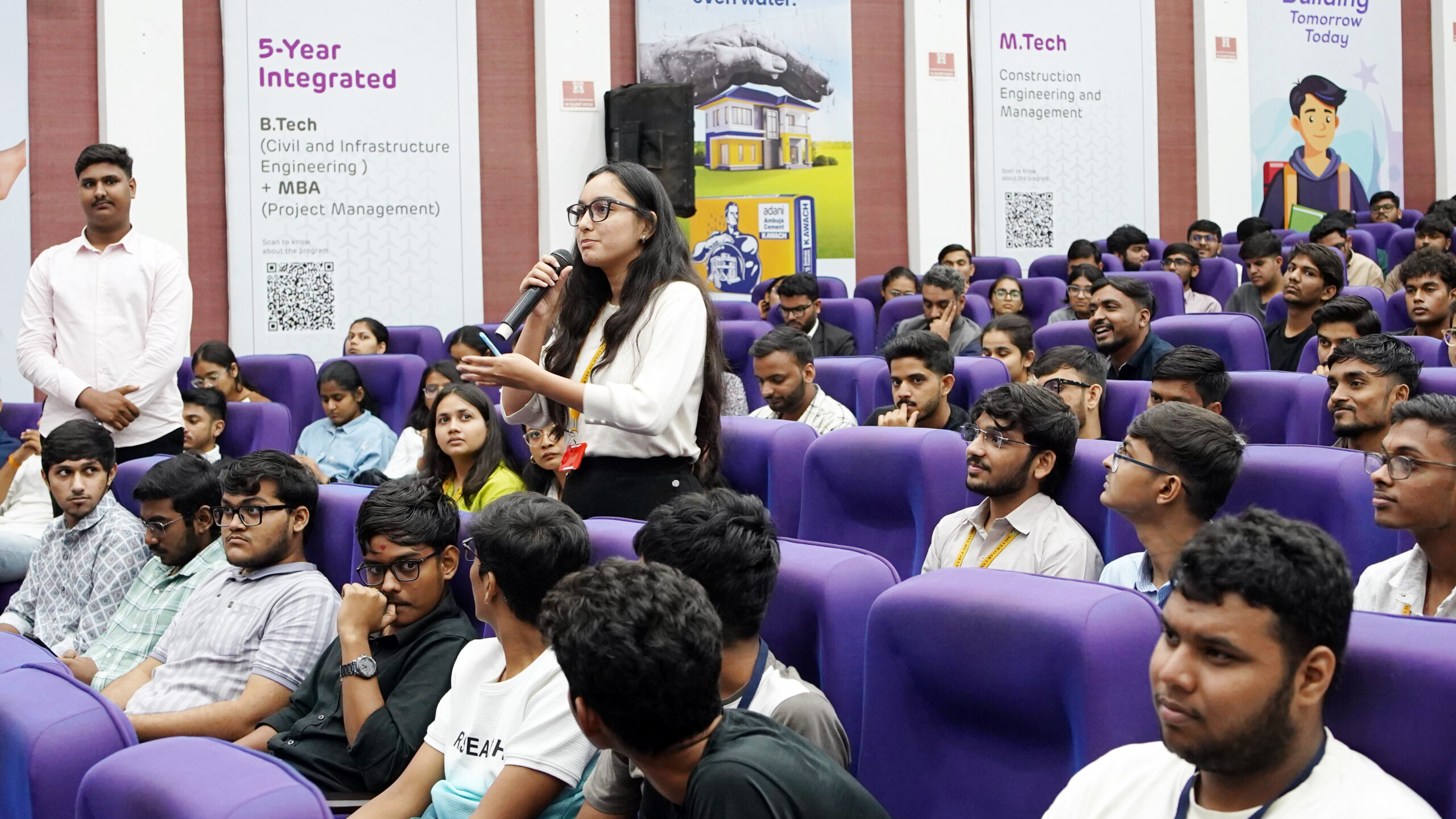A woman speaks into a microphone during a seminar or lecture to a large audience of students.