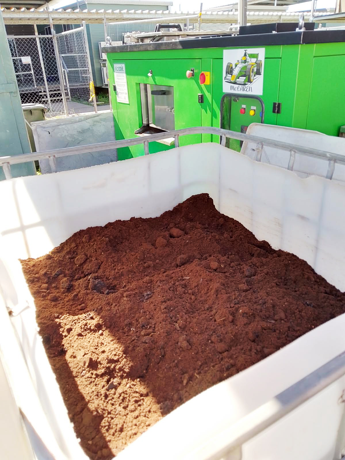 A large bin filled with dark brown compost, with a green industrial composter in the background.