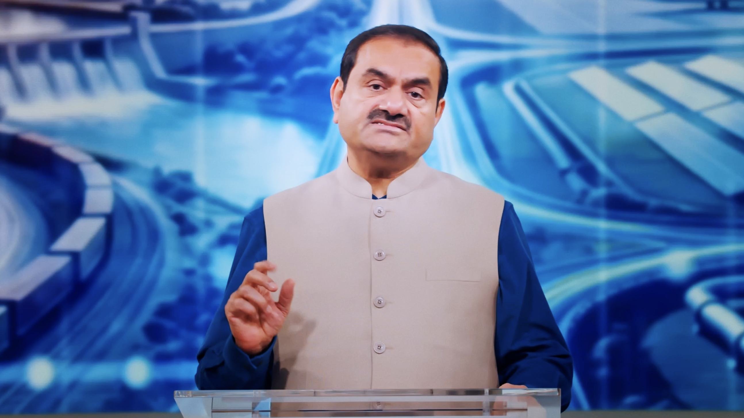 Gautam Adani speaking in a blue shirt and beige waistcoat, against an abstract blue background.