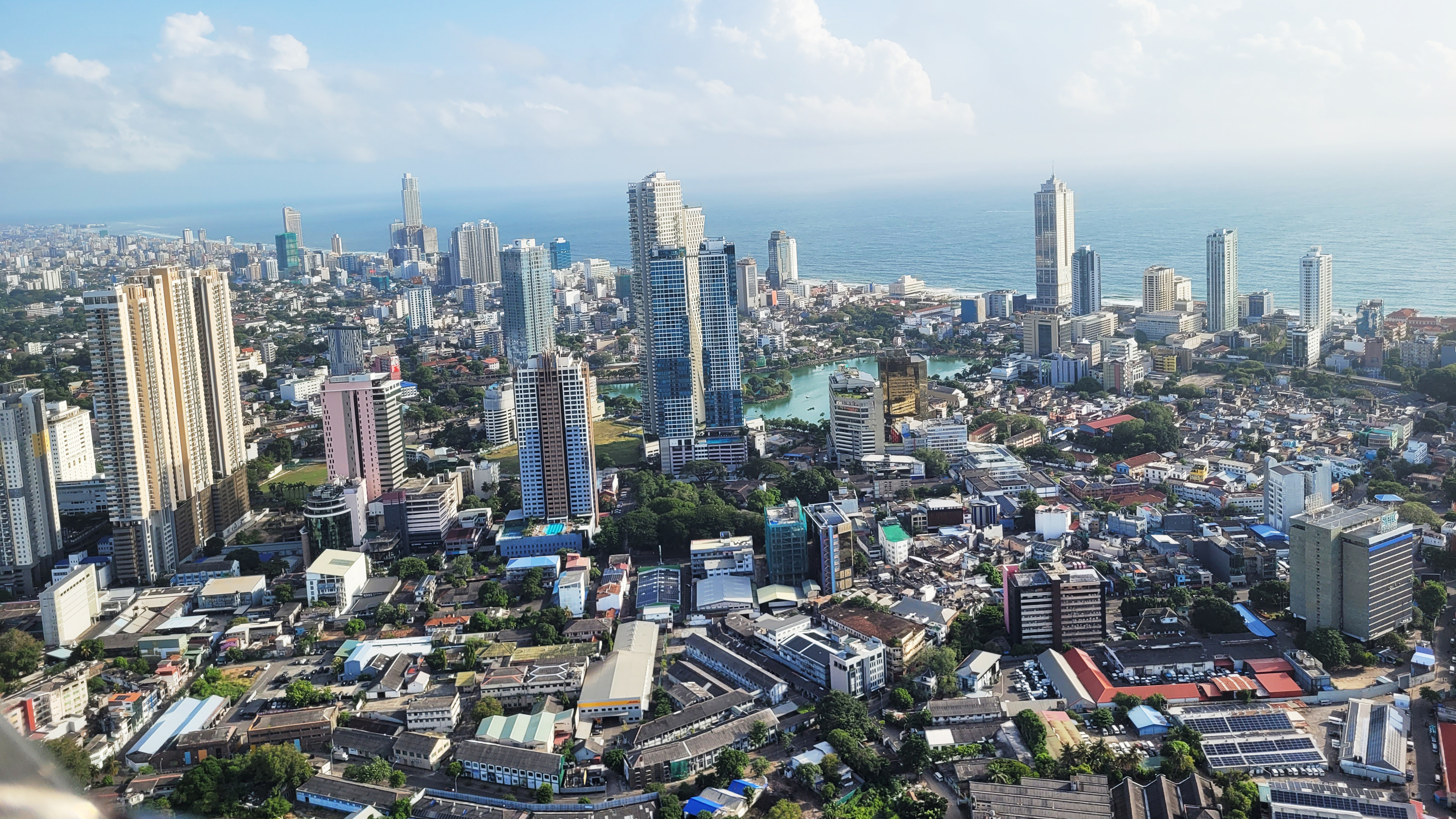 Aerial view of a bustling coastal city with modern skyscrapers, a green lake, and an ocean horizon.