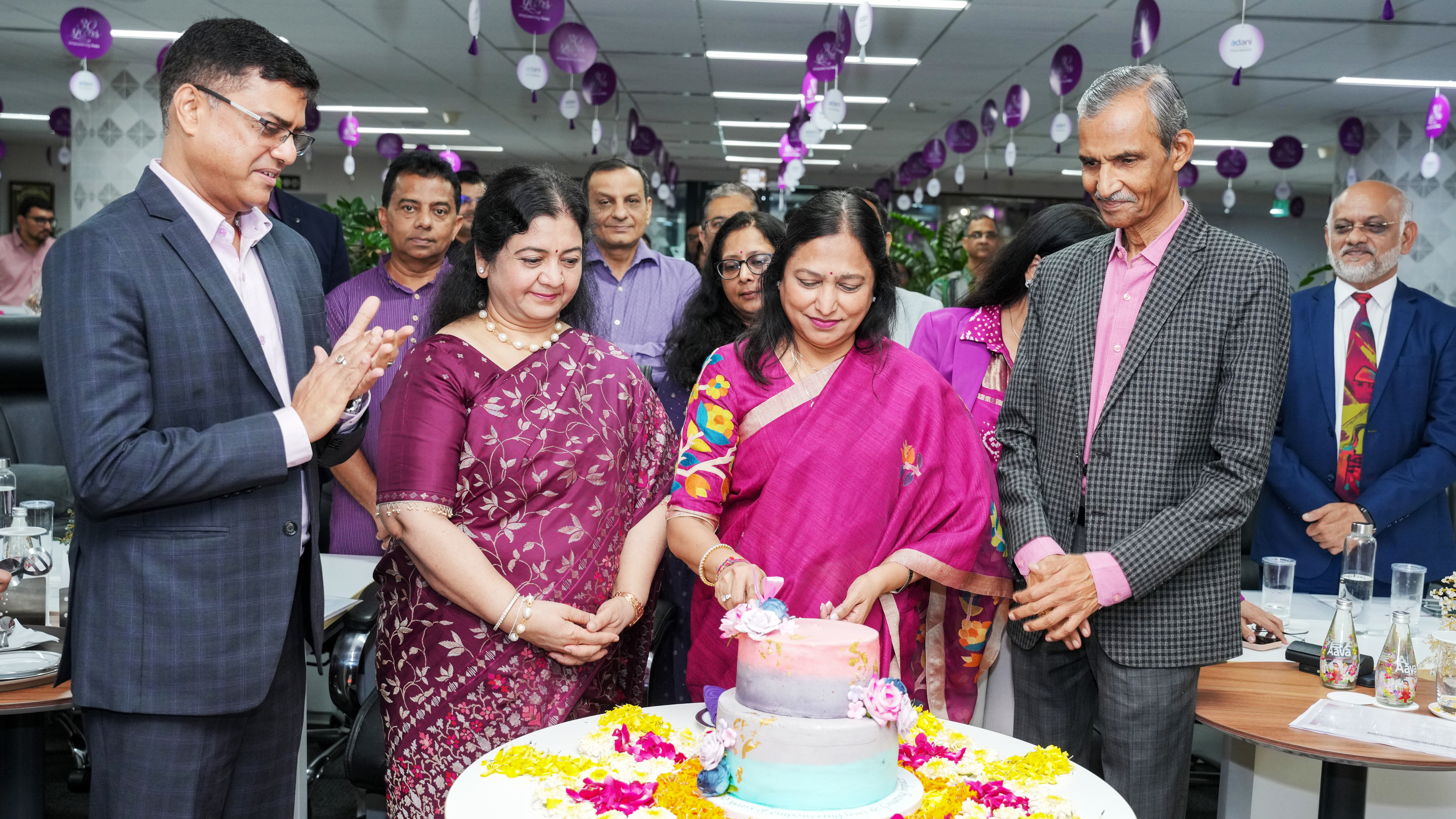 Group celebrates with two women cutting a floral tiered cake, surrounded by applauding guests.