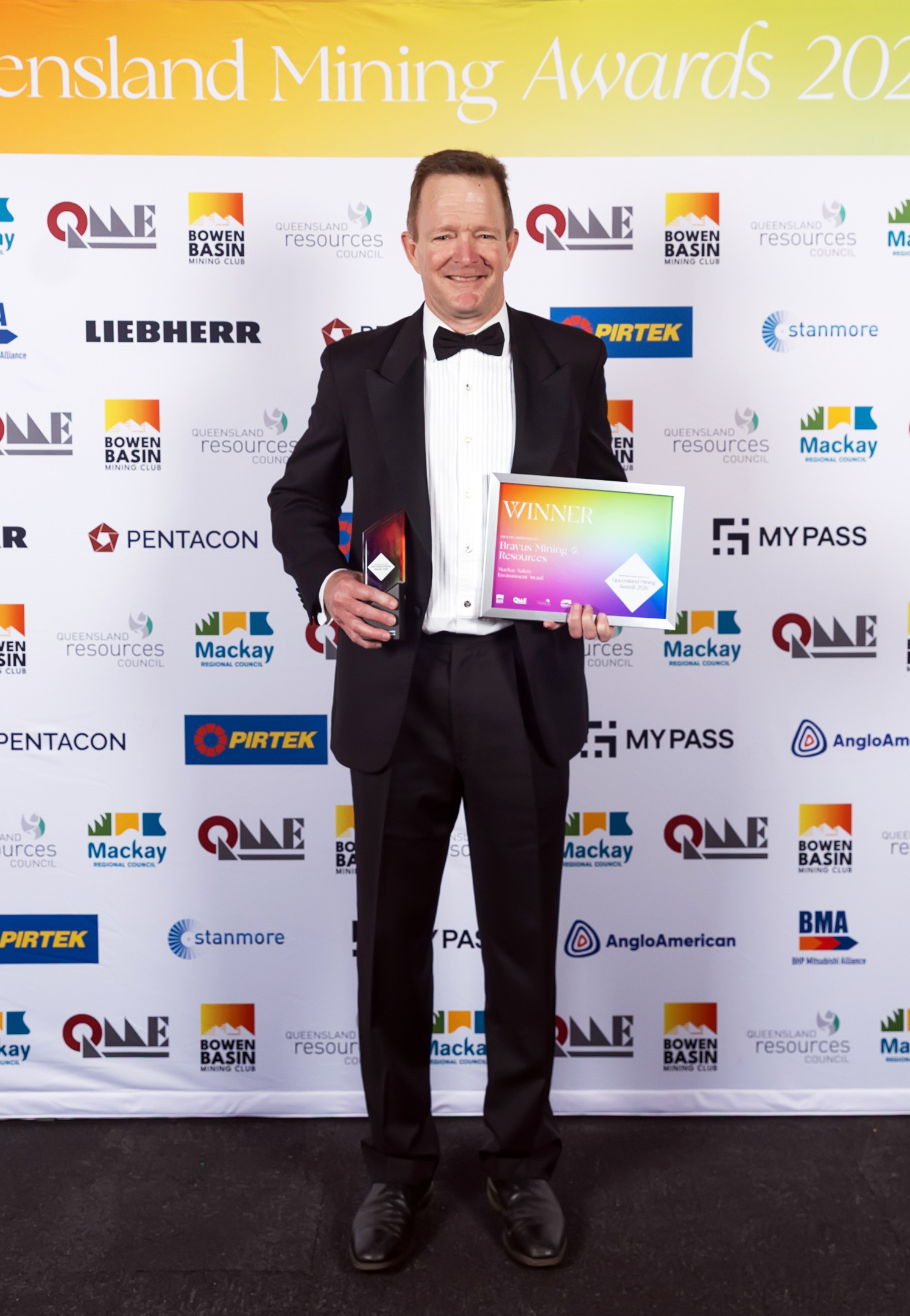 A man in a tuxedo holding two awards at the Queensland Mining Awards event, smiling at the camera.