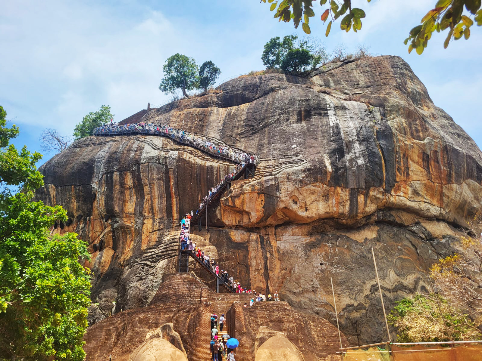 A large rock formation, Sigiriya, with hundreds of people ascending winding stairs towards its summit under a clear sky.