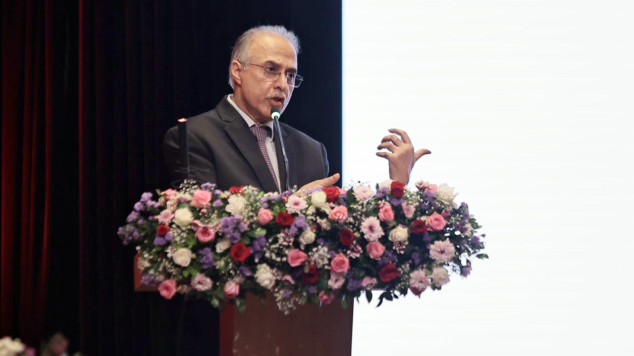 A man in a suit and glasses speaks into a microphone at a podium adorned with colorful flowers.