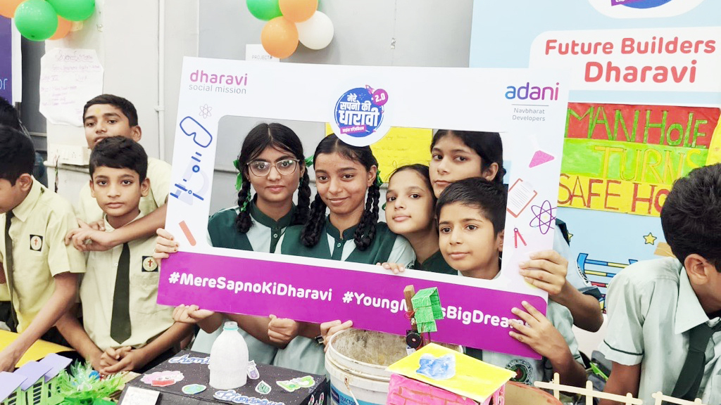 Children in uniform proudly display a "Future Builders Dharavi" photo frame with science projects.
