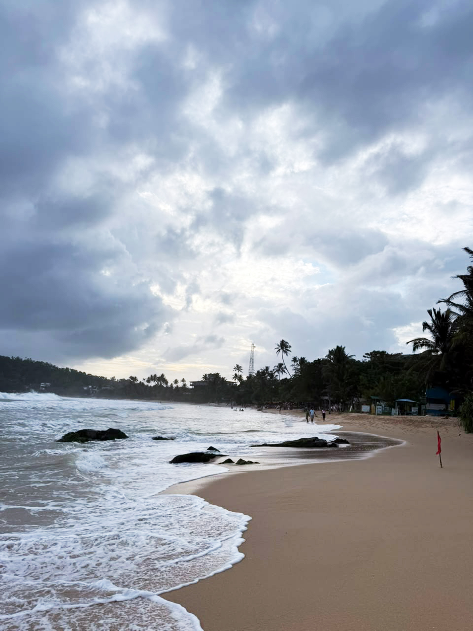Waves crash on a sandy tropical beach under a dramatic cloudy sky, with palm trees and distant people.