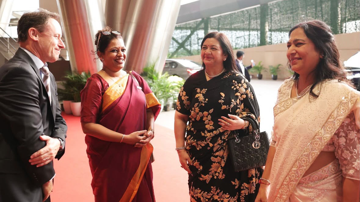 A man in a suit speaks with three smiling women in traditional saris at an event.