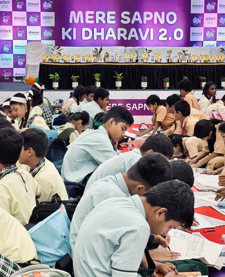 Students drawing on the floor at a "MERE SAPNO KI DHARAVI 2.0" event, focusing on their creative work.