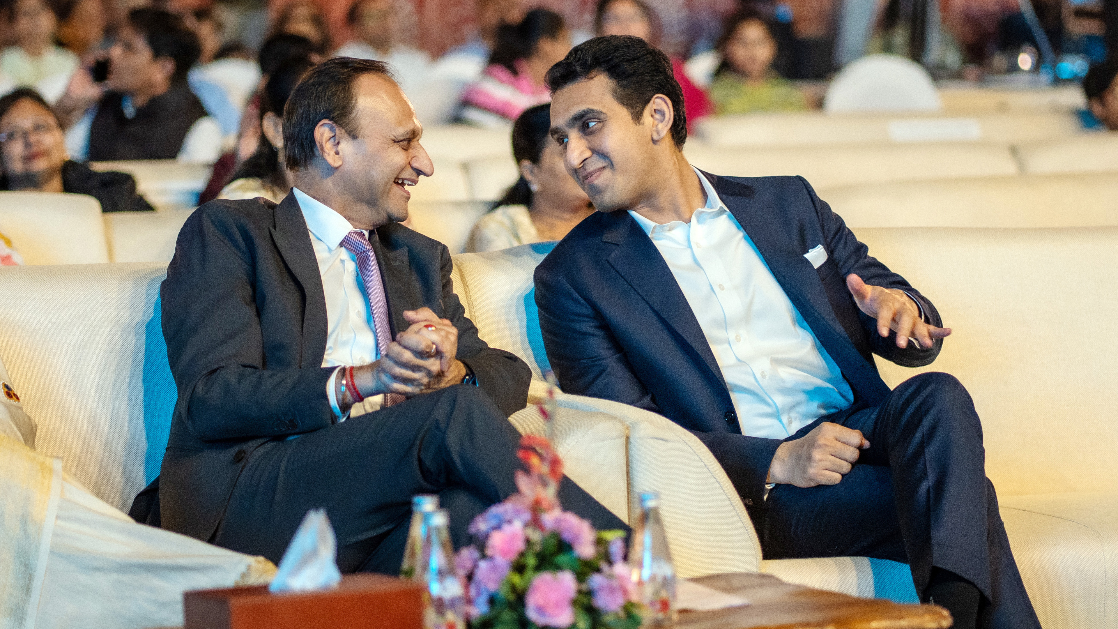 Two men in suits sitting on a couch, talking and smiling at an event.