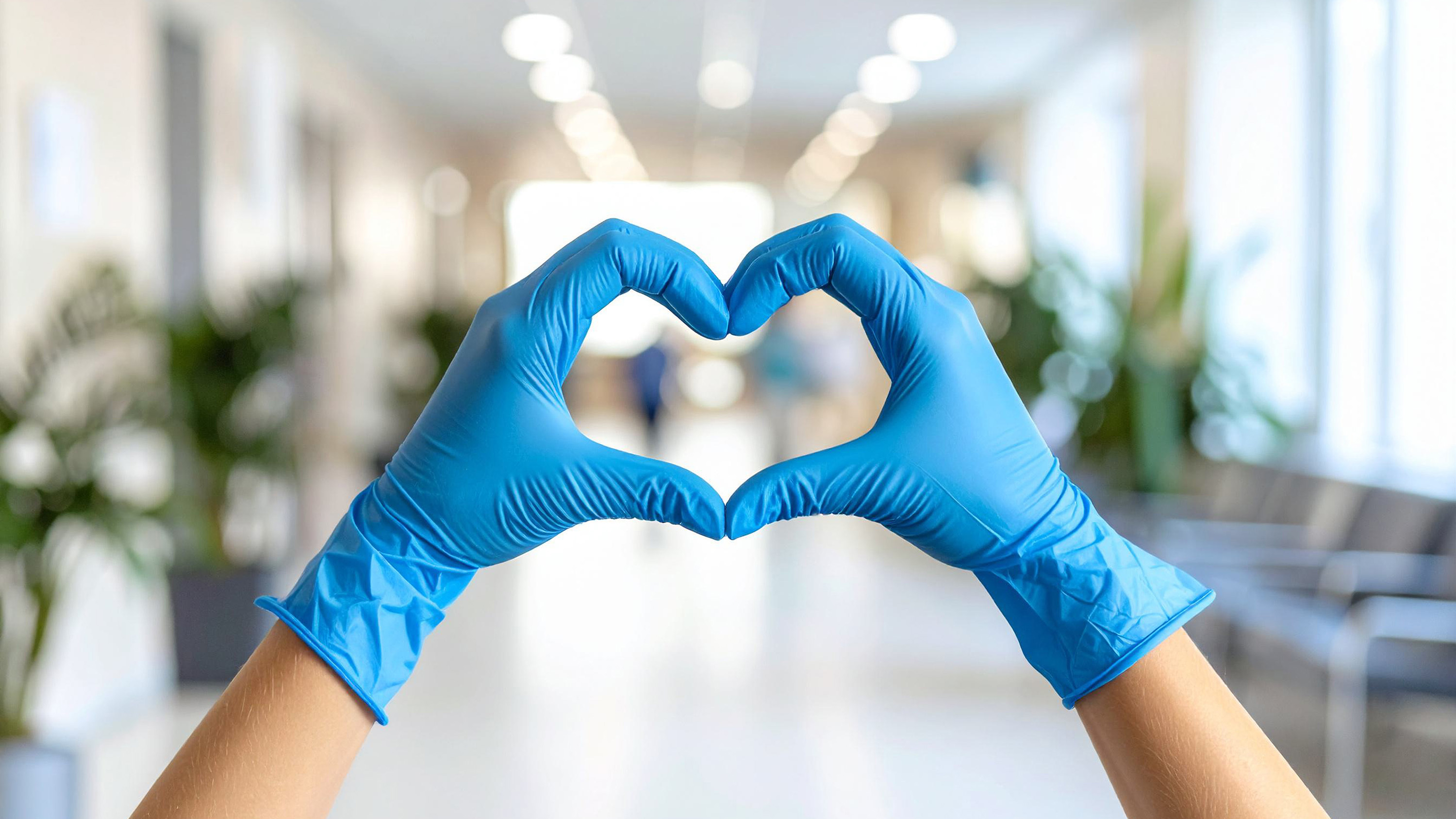 Hands in blue medical gloves forming a heart shape, symbolizing care and health, in a hospital corridor.