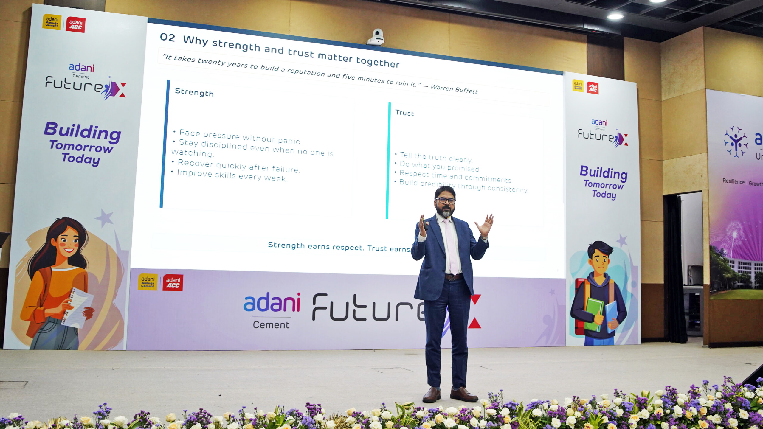 A man in a suit presents on stage about strength and trust, with a large screen displaying the topic and key points.