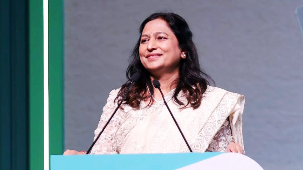 A smiling woman with dark hair and a patterned sari speaks at a podium with microphones.