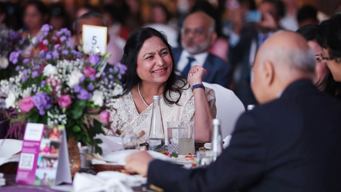 A smiling woman in a white saree at a formal event, engaging in conversation at a beautifully set table.