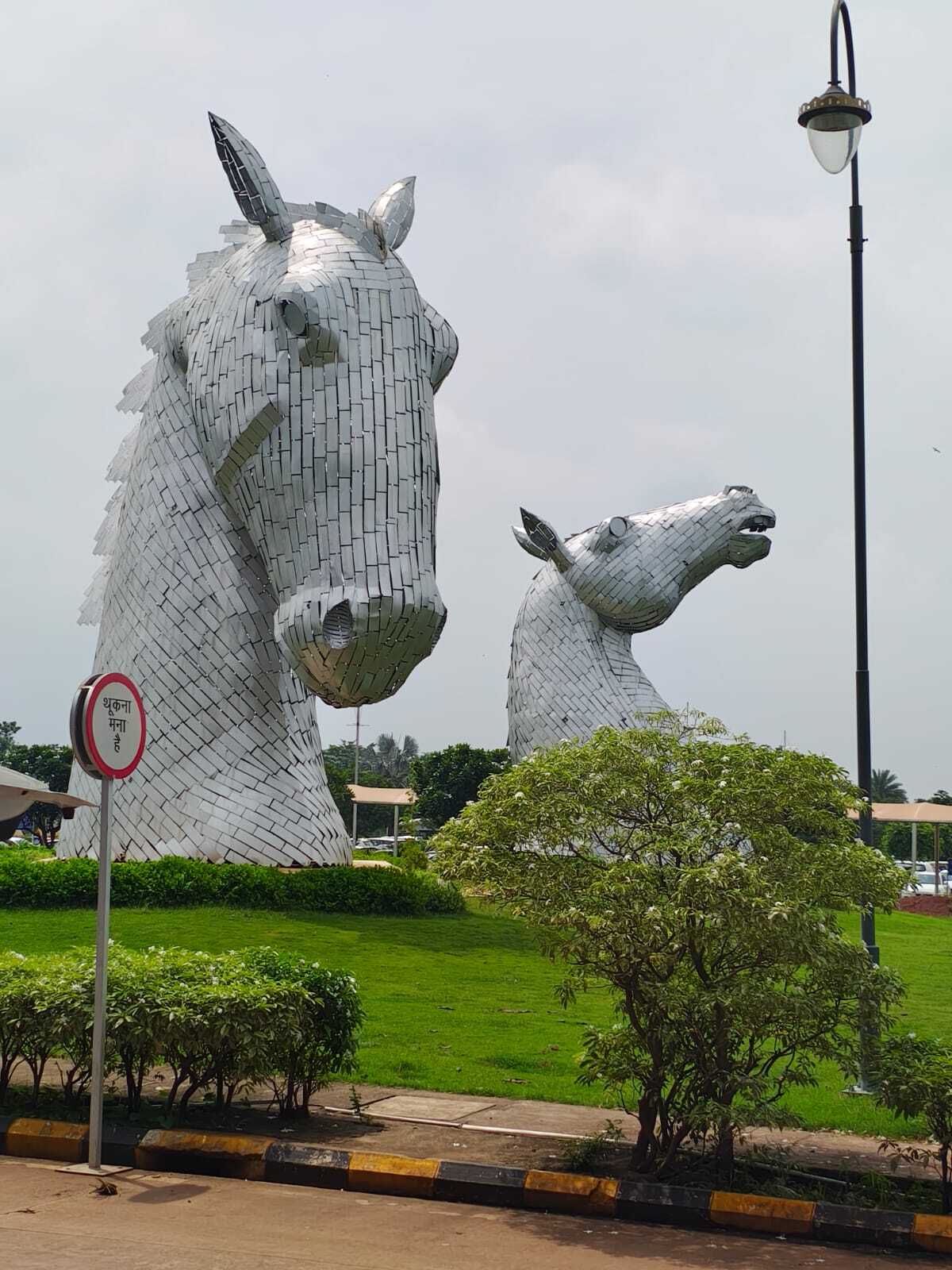 Two large, modern horse head sculptures made of silver-colored metal plates in a green park setting.