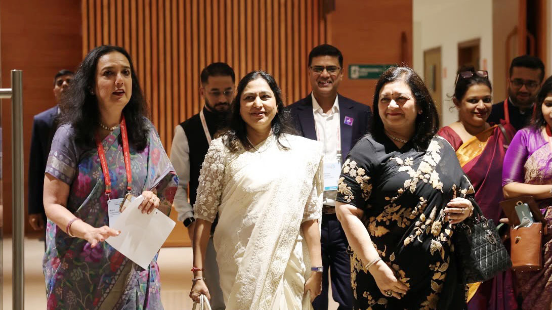 Three women in traditional Indian attire walk through a modern building, smiling, with others in the background.