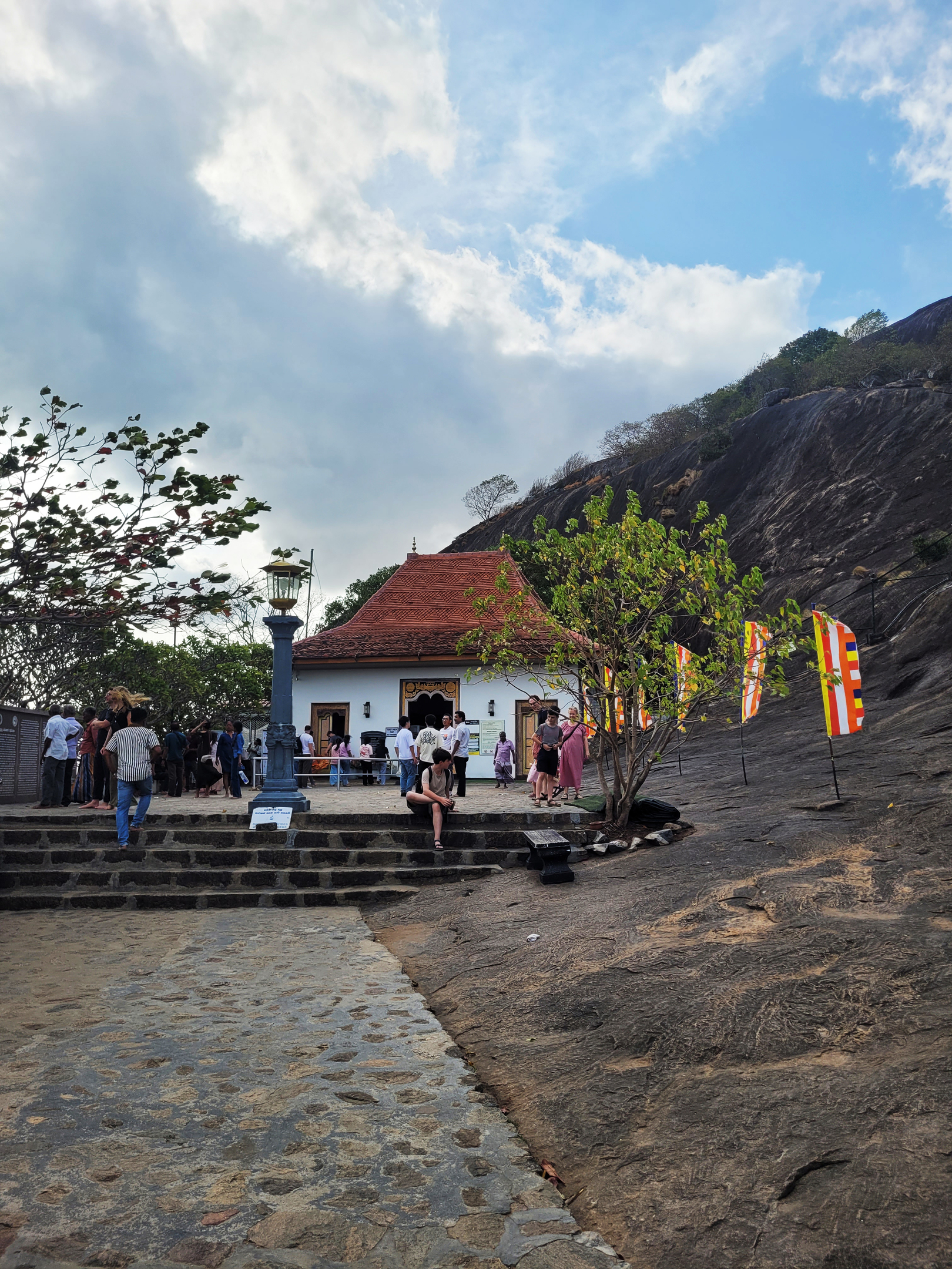 A white temple with a red roof nestled into a rocky hillside, with people gathered on stone steps.